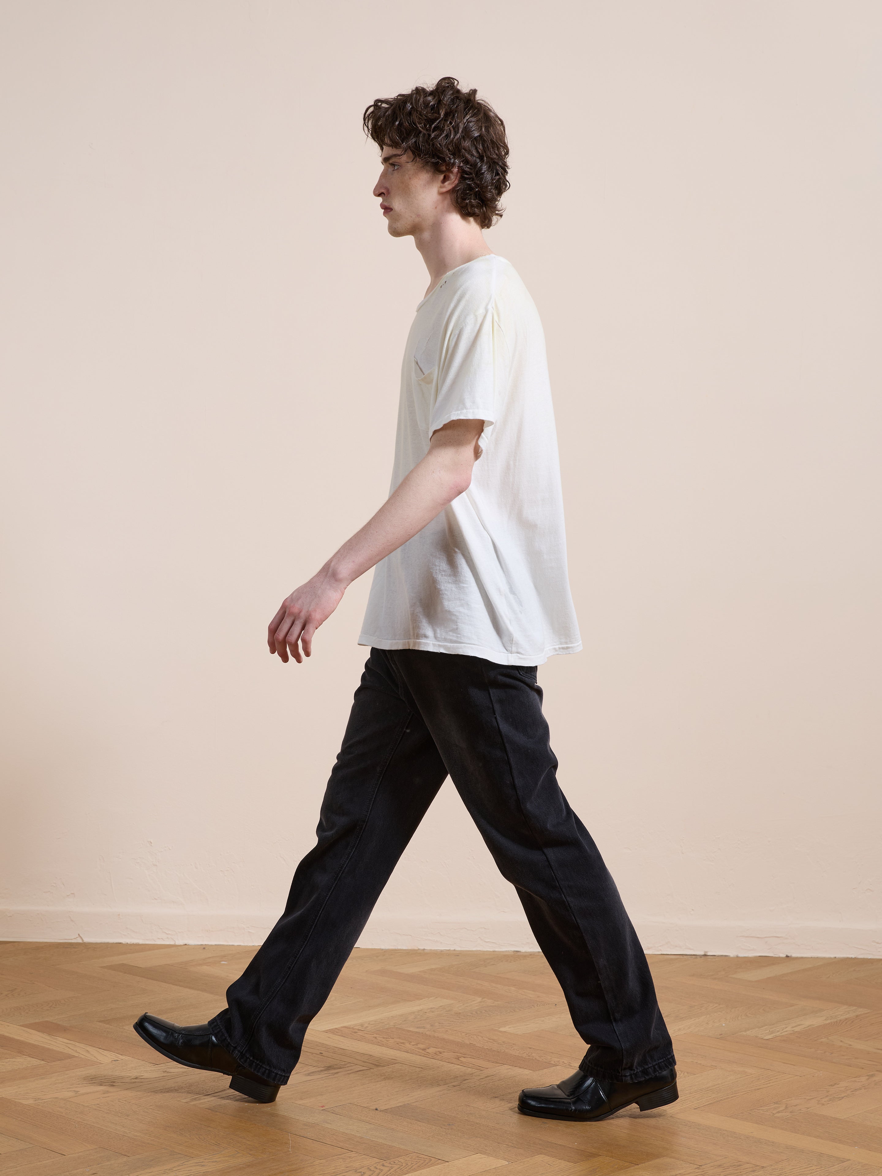 A young man with curly hair wears the FOUND Weathered Pocket Tee, black jeans, and black shoes as he walks indoors on a parquet floor beside a plain beige wall.