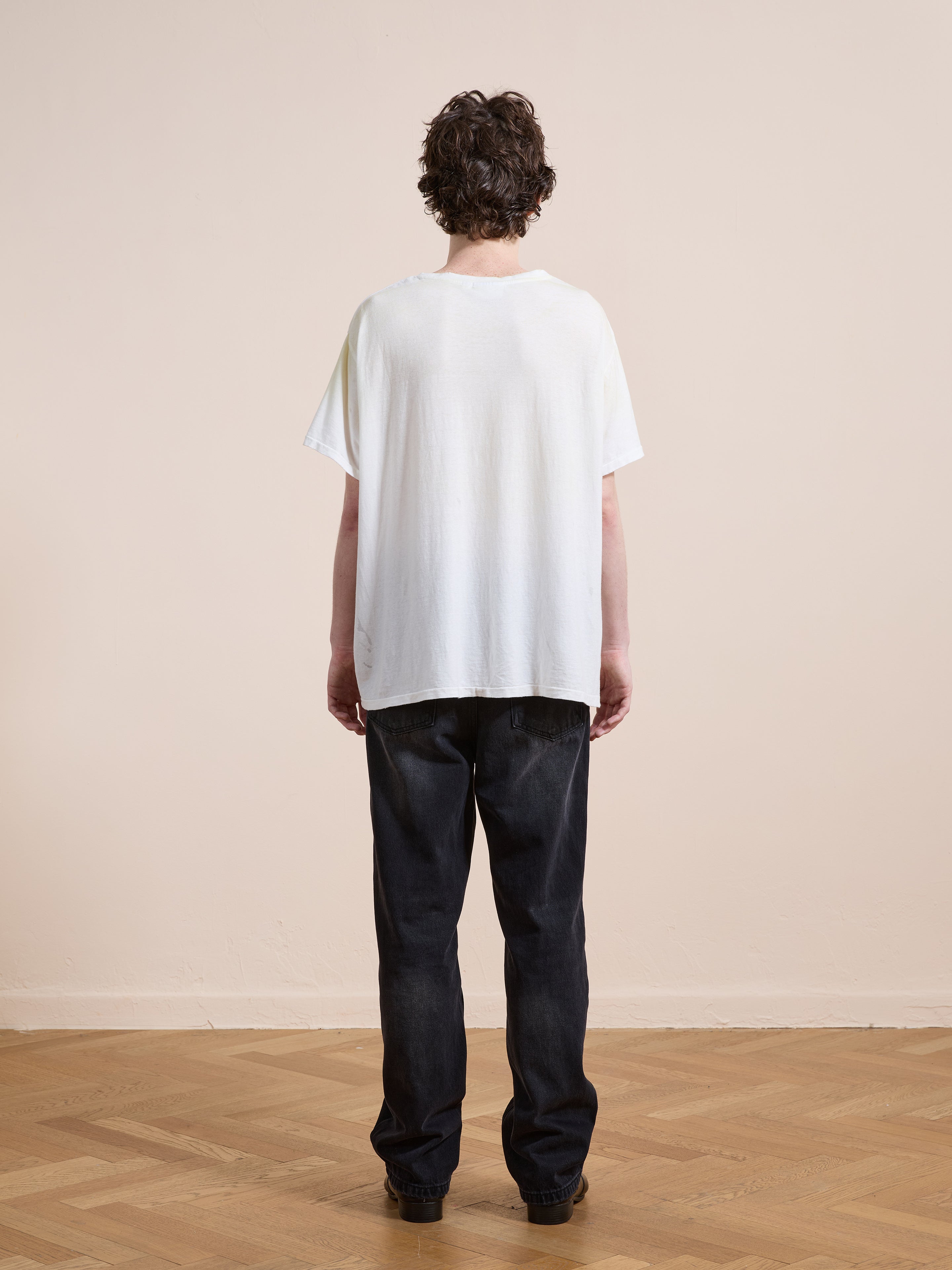 A person with short curly hair stands facing away, wearing the FOUND Weathered Pocket Tee in white with black jeans on a wooden floor against a plain beige wall.