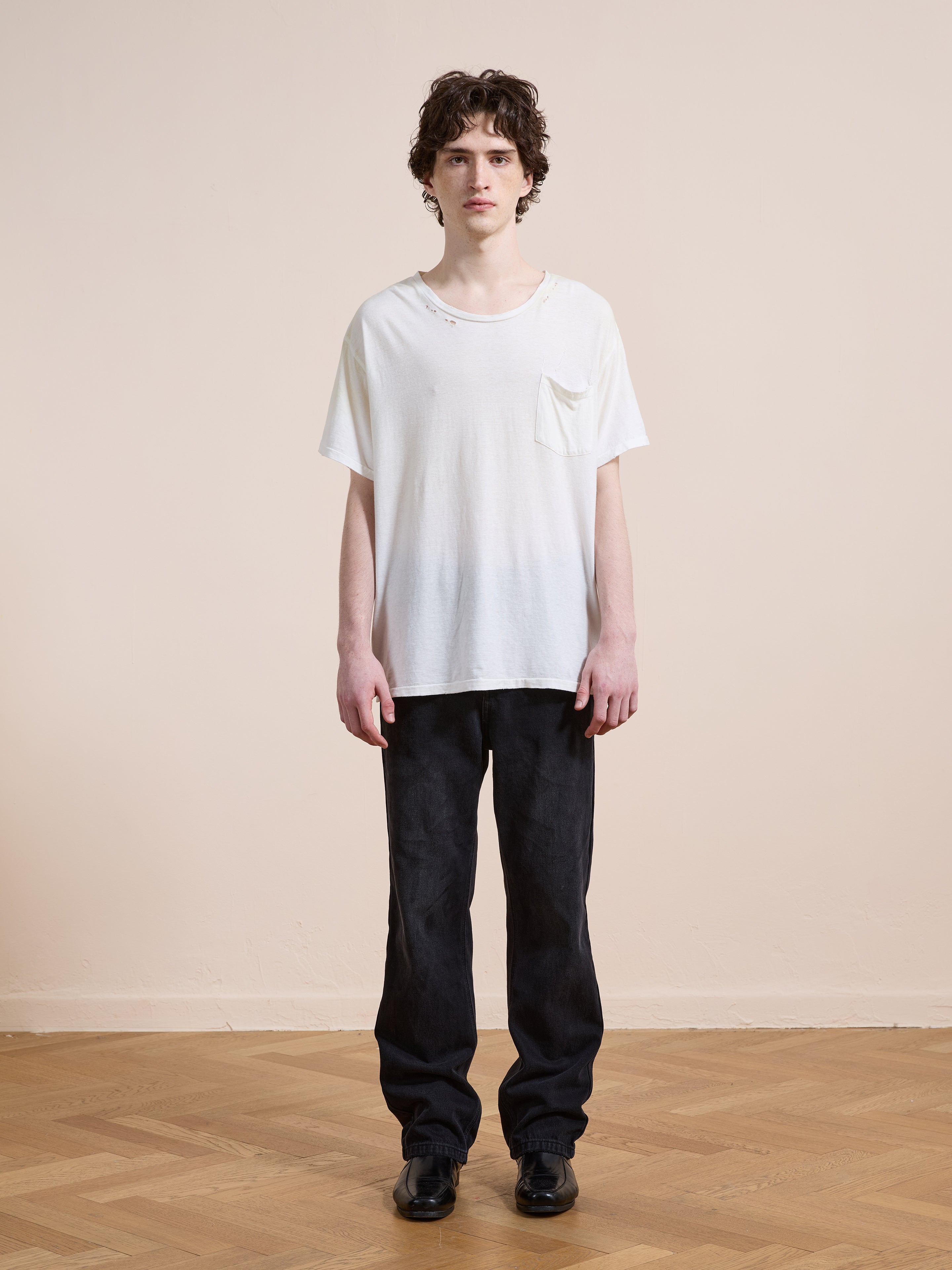 A person with short curly hair stands on a wooden floor, wearing the FOUND Weathered Pocket Tee, black pants, and black shoes, against a plain beige wall.
