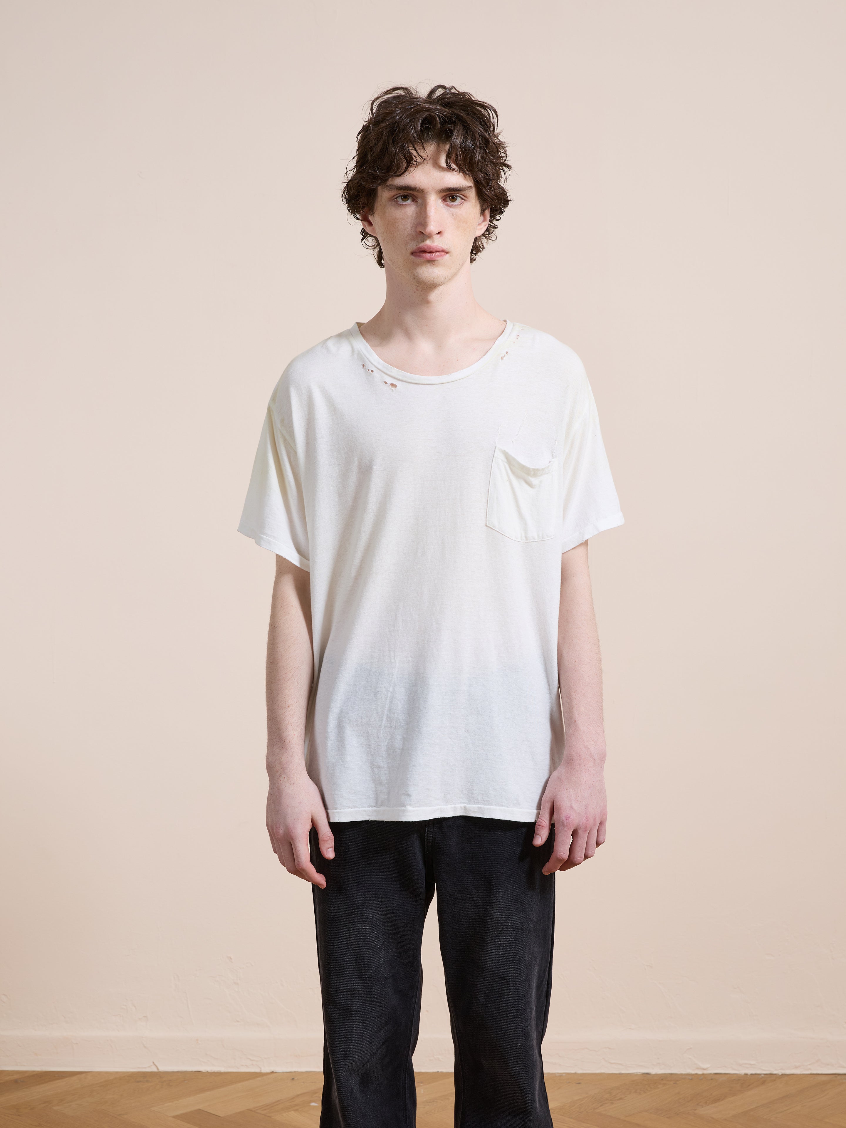 A person with medium-length curly hair wears the FOUND Weathered Pocket Tee, a relaxed fit unisex white shirt with a chest pocket, paired with black pants, standing indoors against a plain light-colored wall.