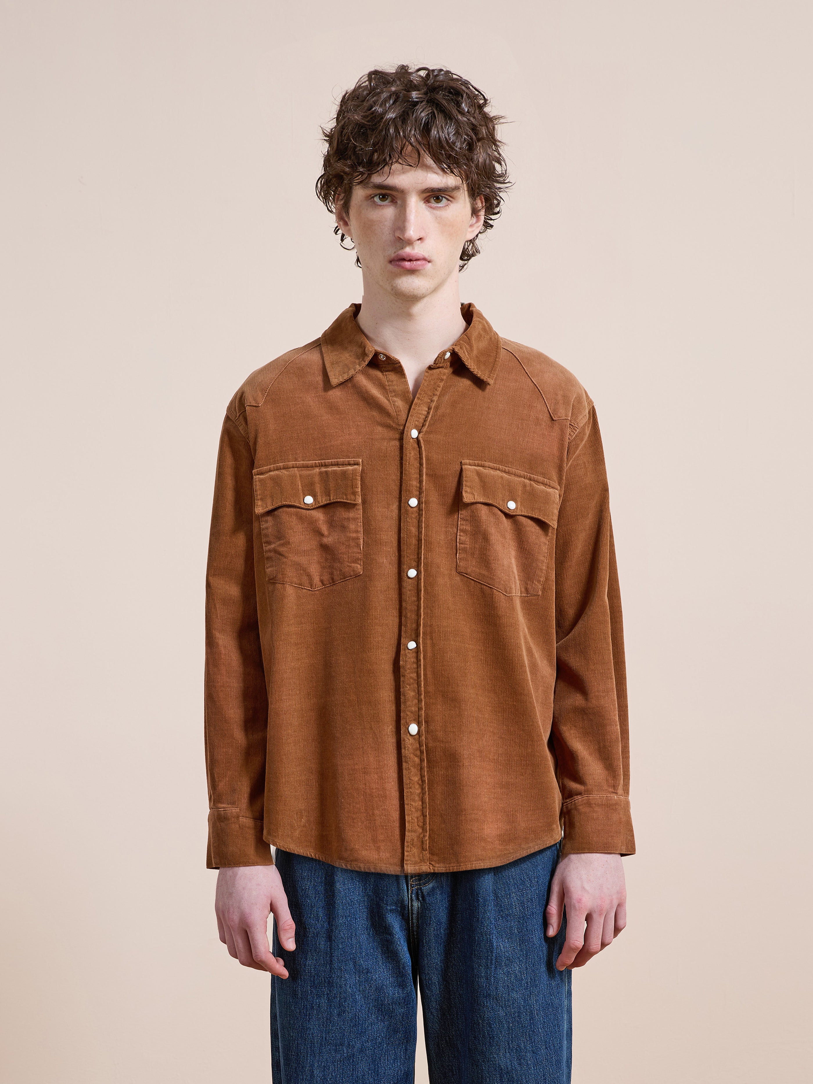 A person with curly hair wears the FOUND Western Corduroy Shirt, featuring two chest pockets and a relaxed unisex fit, paired with blue jeans against a plain beige background.