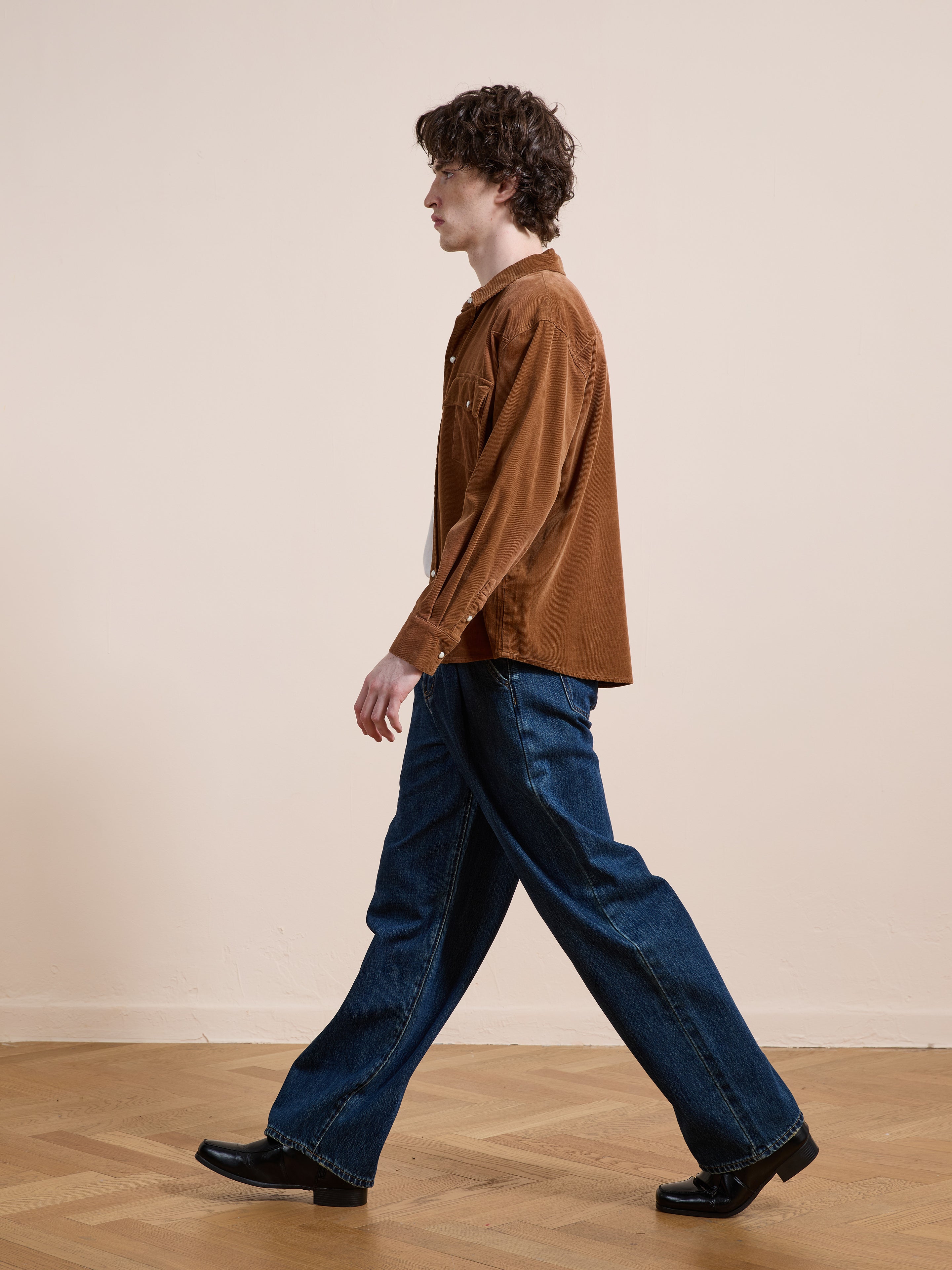 A person with curly hair walks indoors wearing the FOUND Western Corduroy Shirt, paired with blue wide-leg jeans and black shoes. The background is a plain light wall and wooden floor.
