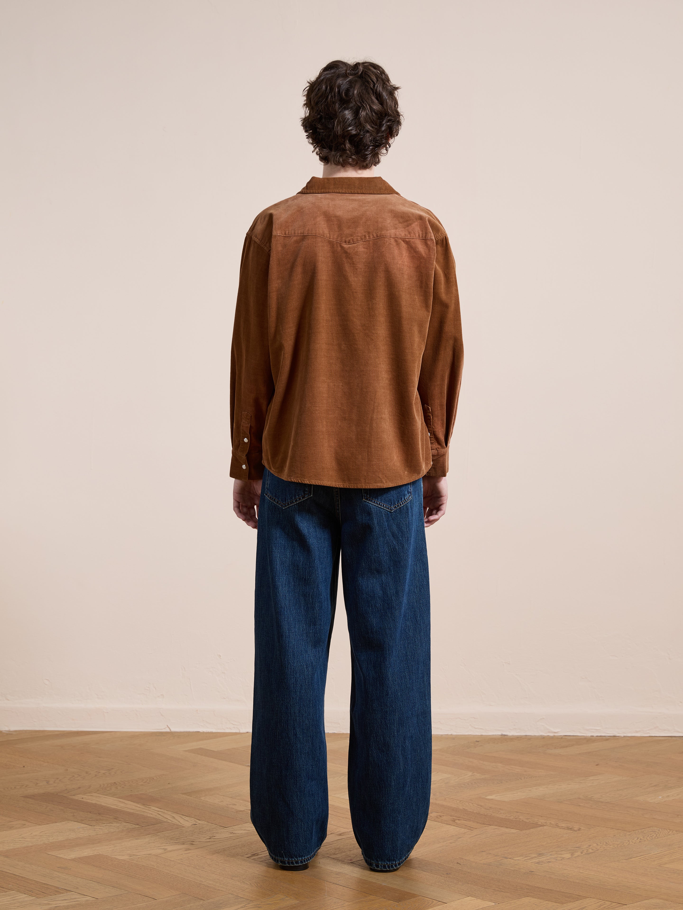A person stands indoors facing away from the camera, wearing the FOUND Western Corduroy Shirt and loose blue jeans on a wooden floor with a plain light-colored wall in the background.
