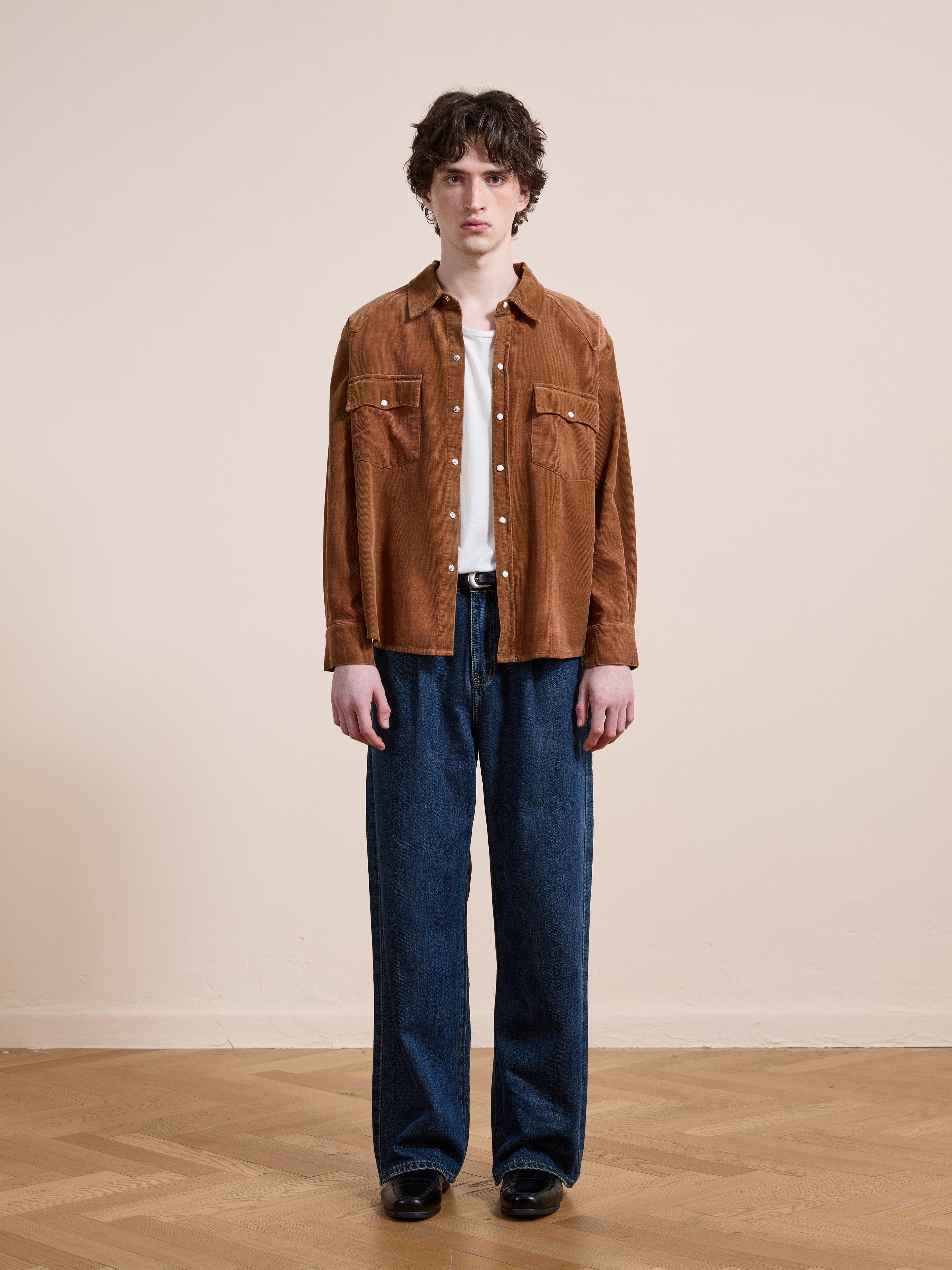 A person stands indoors on a wooden floor, facing the camera against a plain beige wall, wearing the FOUND Western Corduroy Shirt over a white t-shirt, paired with loose blue jeans and black shoes.