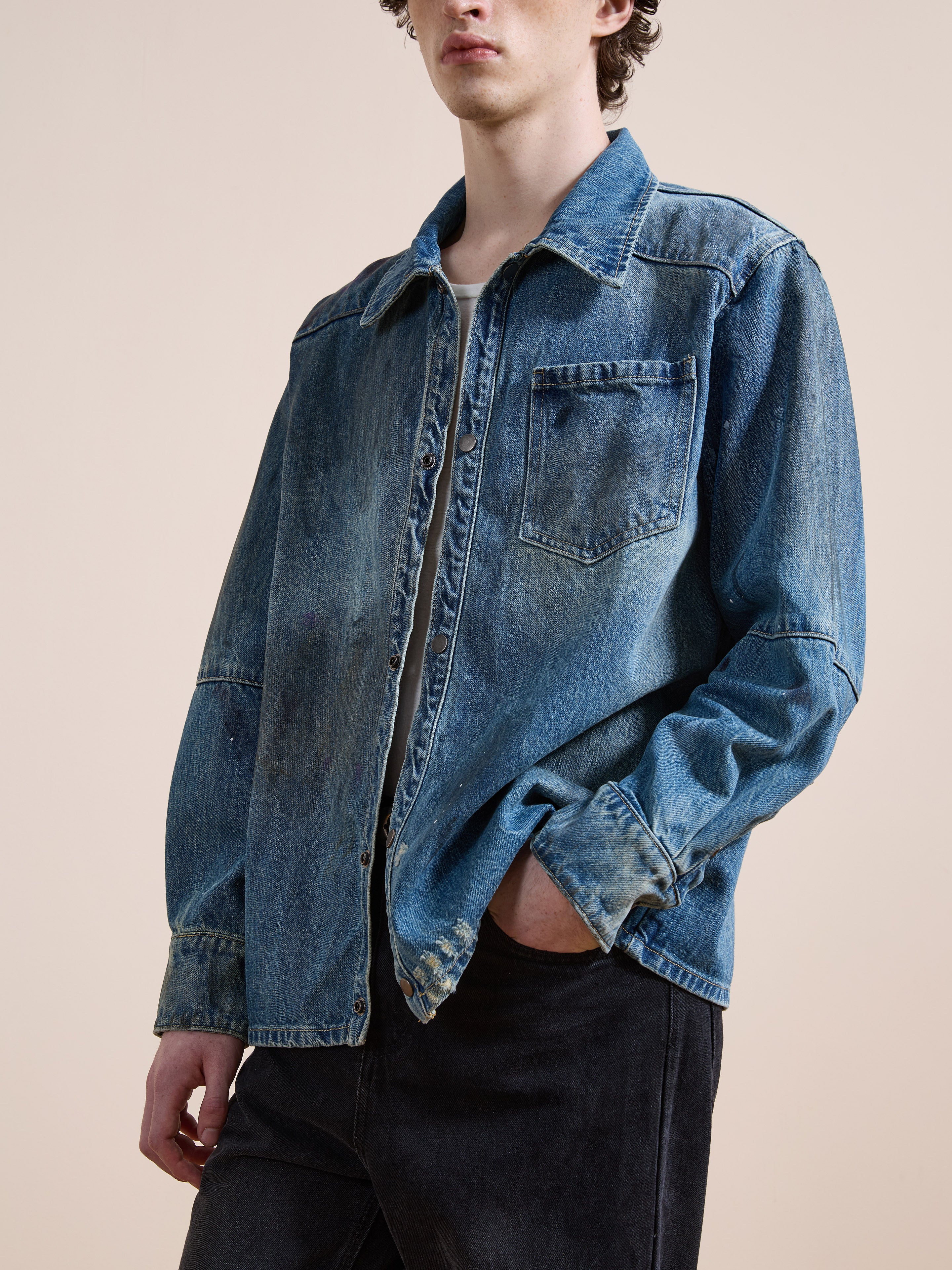 A person in the FOUND Western Denim Painter Shirt, worn loose over a white tee and black jeans, stands with one hand in their pocket against a plain background.