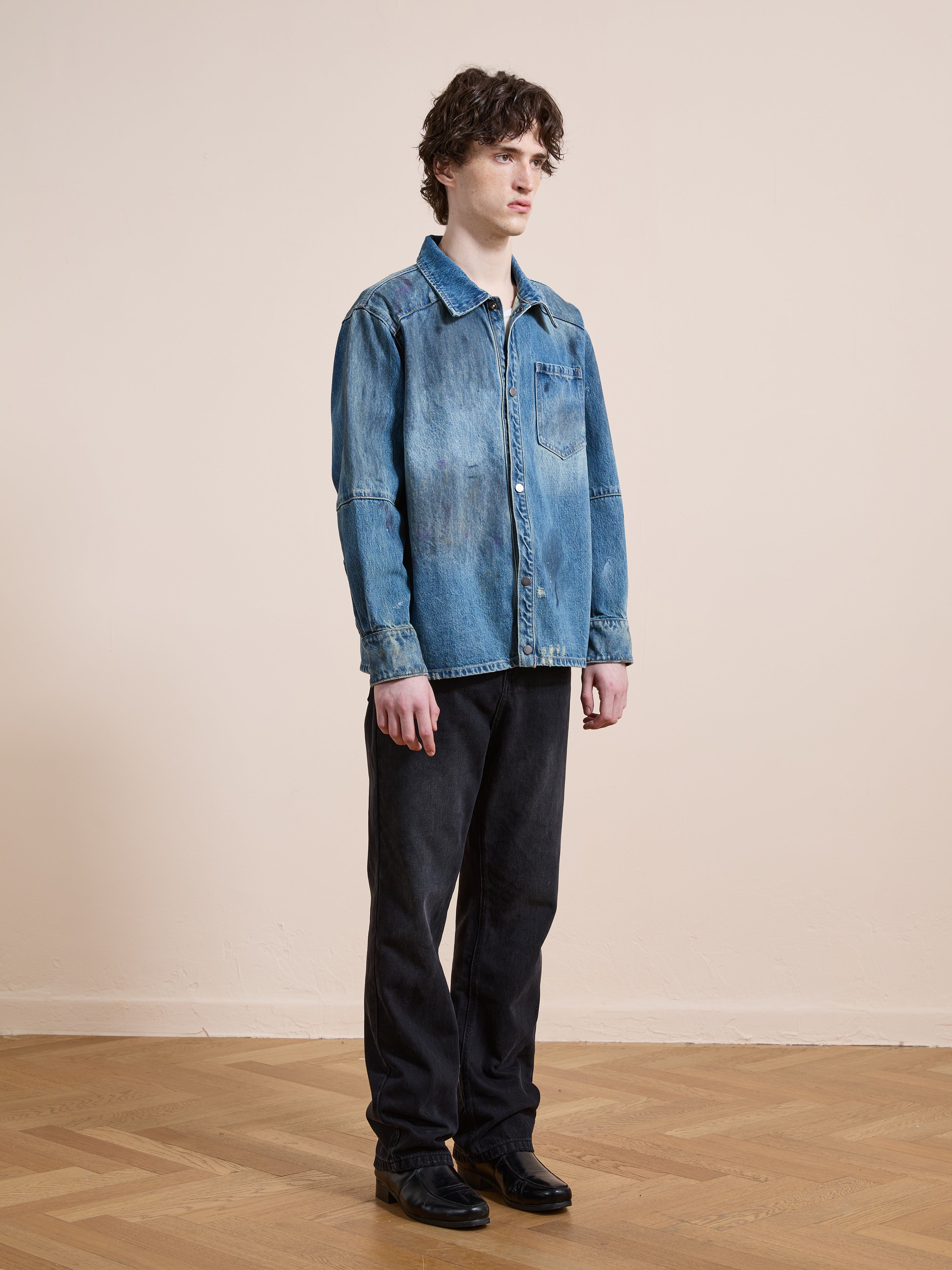 Indoors on a wooden floor, someone faces sideways by a beige wall, wearing the FOUND Western Denim Painter Shirt in vintage blue, paired with black pants and shoes.