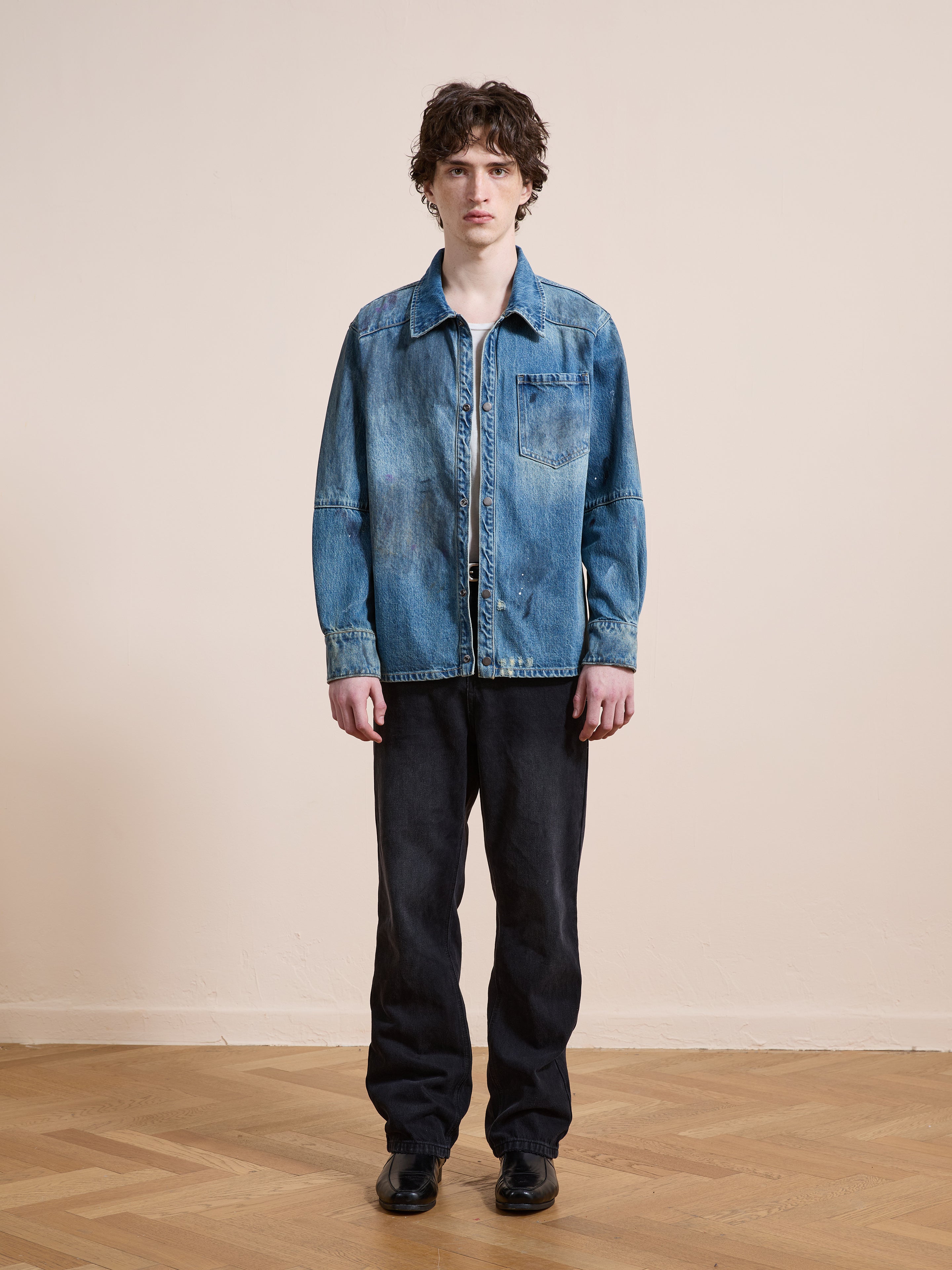 A person wearing the FOUND Western Denim Painter Shirt stands indoors on a wooden floor, paired with black jeans and black shoes, facing the camera against a plain beige wall.