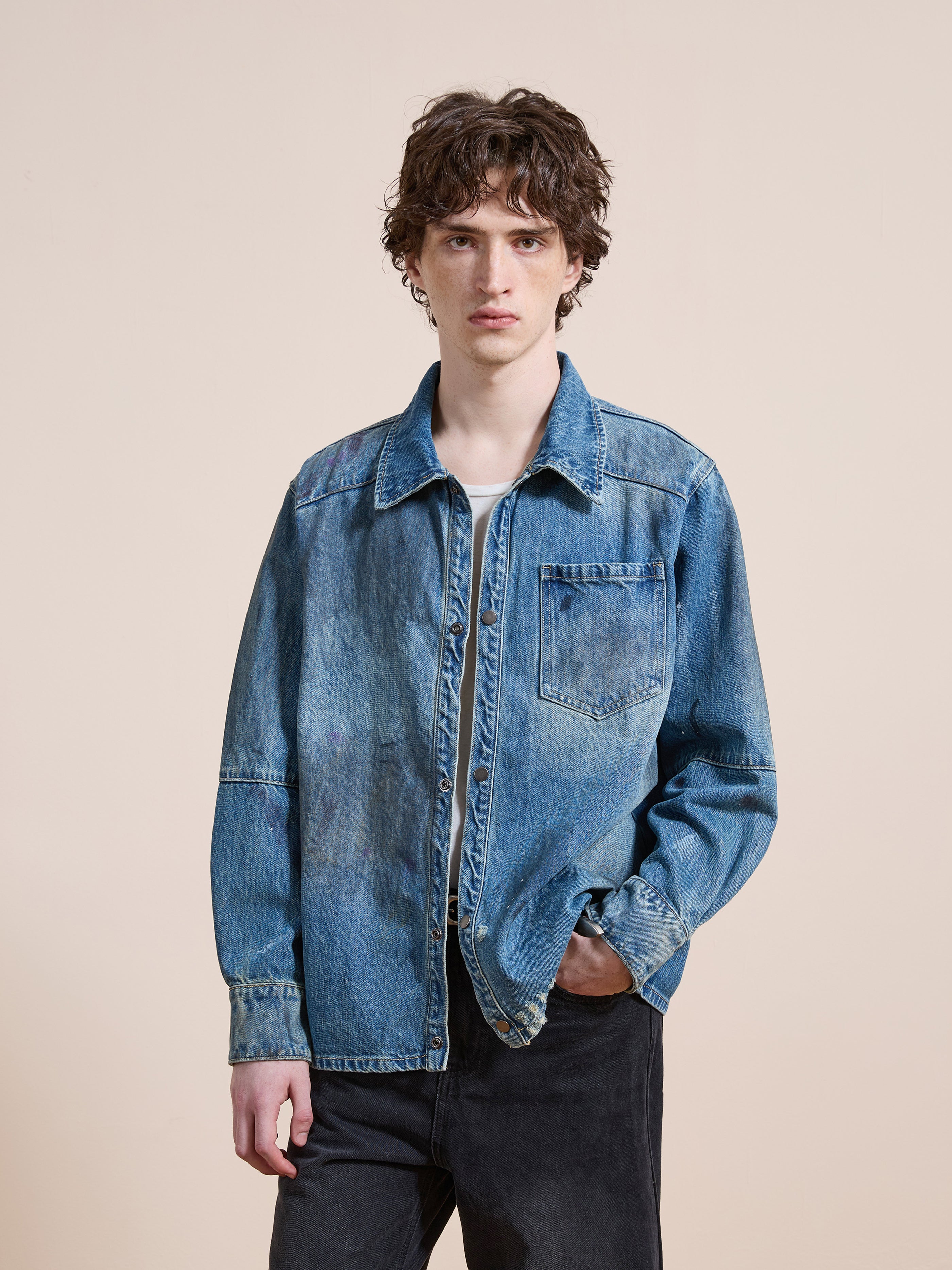 A person models the FOUND Western Denim Painter Shirt layered over a white tee and black jeans, standing against a plain beige background.
