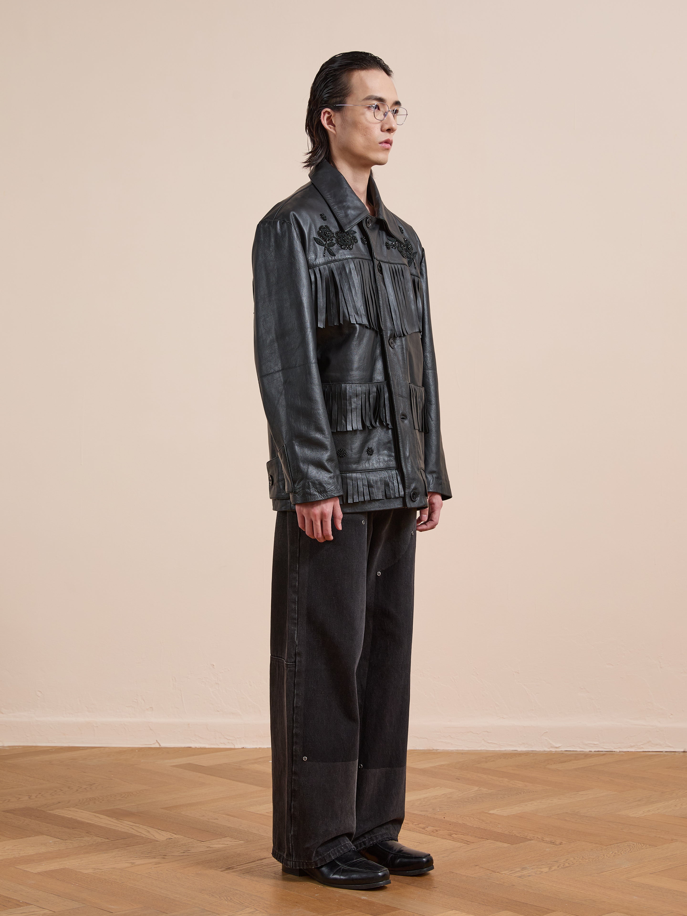 Indoors on a wooden floor, a person in glasses faces left against a beige wall wearing FOUND's Hand Embroidered Leather Fringe Jacket with wide-leg black pants and black shoes.