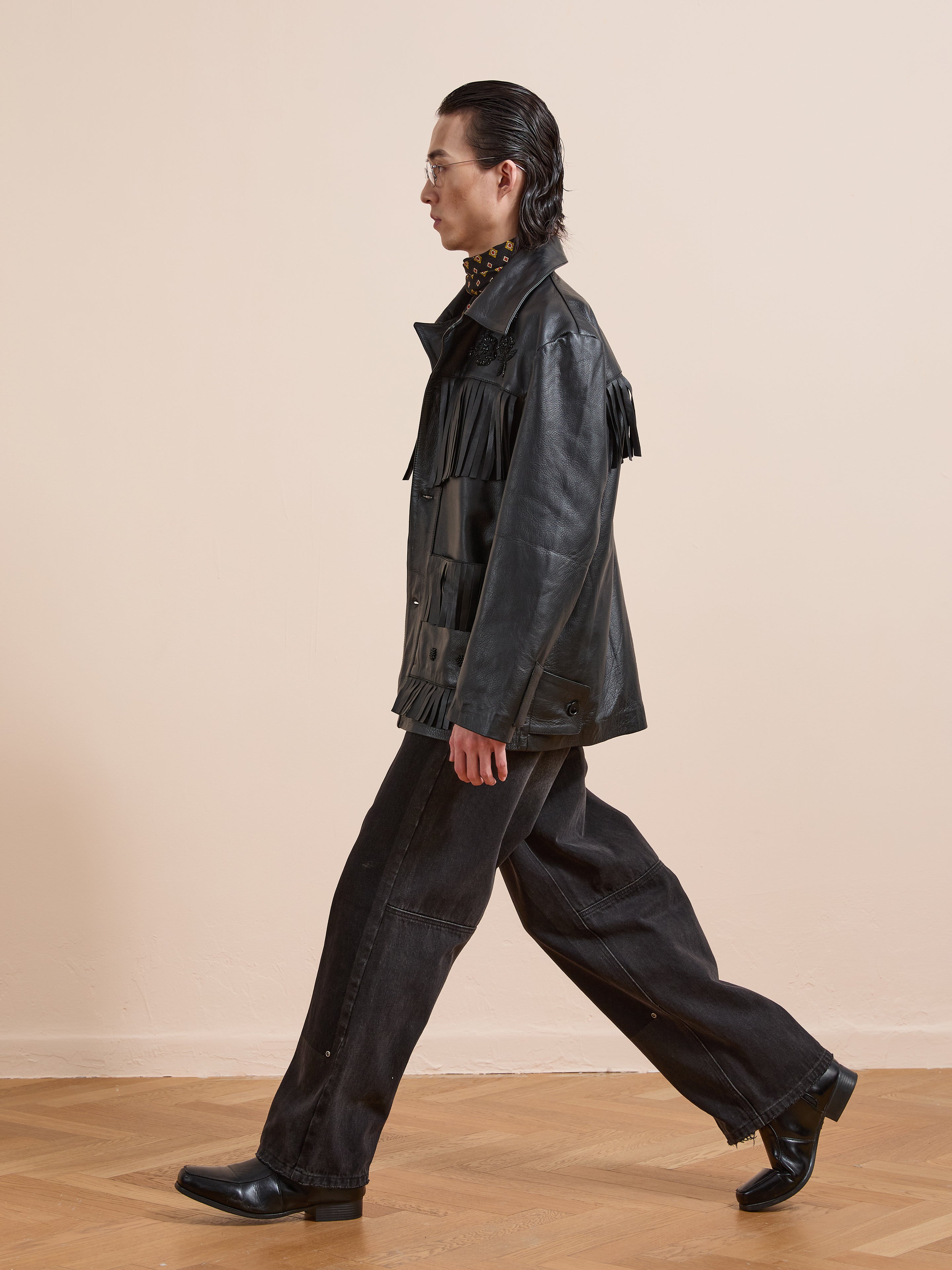 A person in profile walks indoors against a beige background, wearing black flared pants, heeled boots, glasses, and the FOUND Hand Embroidered Leather Fringe Jacket made from genuine cowhide.