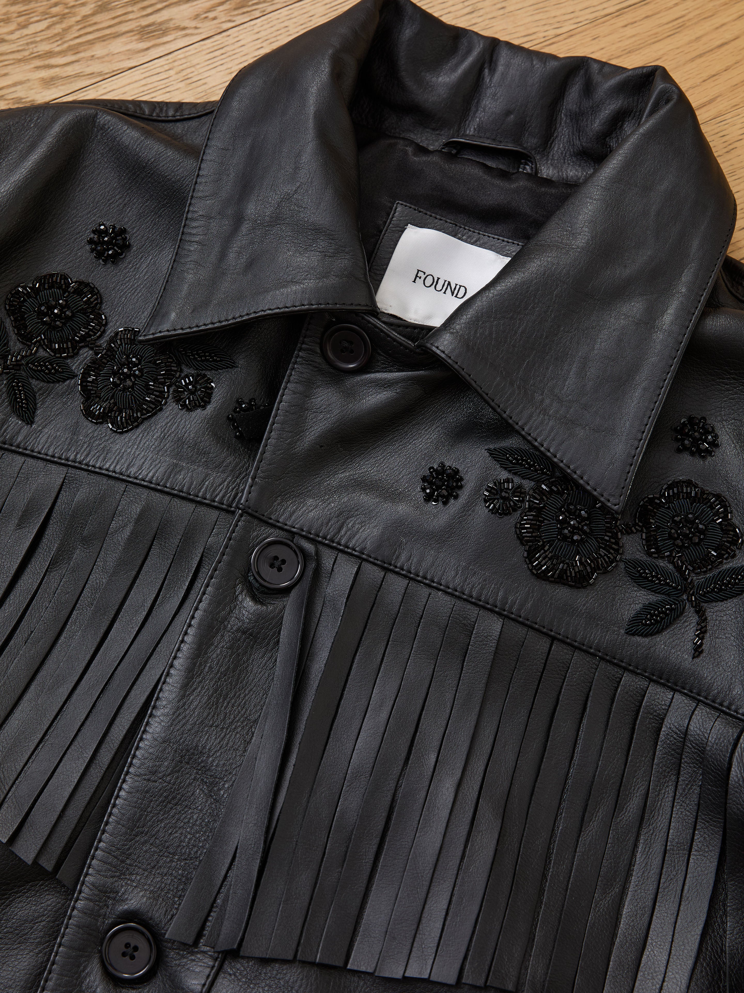 The FOUND Hand Embroidered Leather Fringe Jacket, made from black genuine cowhide with pleated details and beaded floral embroidery, is displayed flat on a wooden surface with its brand label visible.