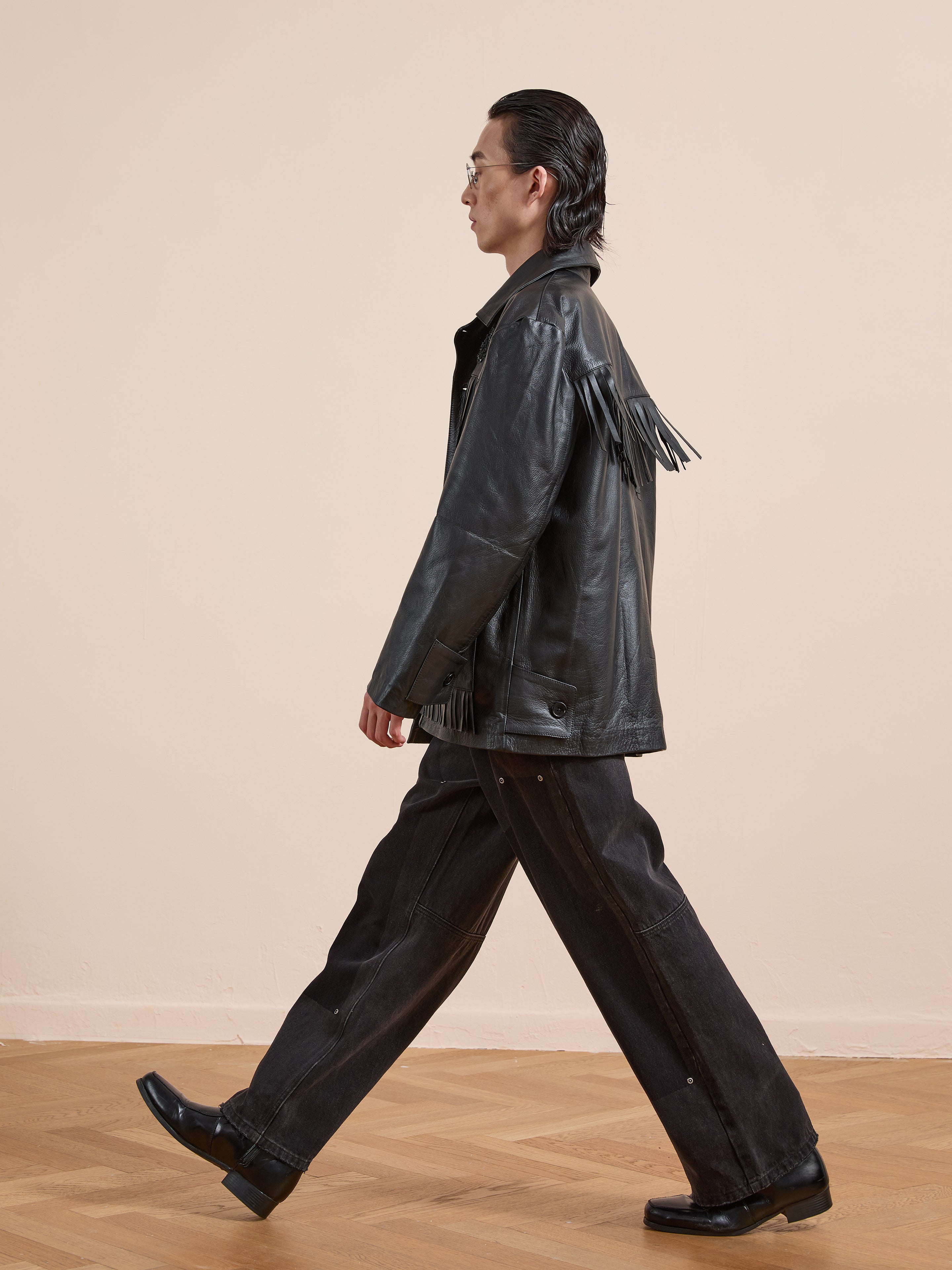 A person in the FOUND Hand Embroidered Leather Fringe Jacket, paired with dark wide-leg jeans and black shoes, walks indoors on a wooden floor against a plain light background.