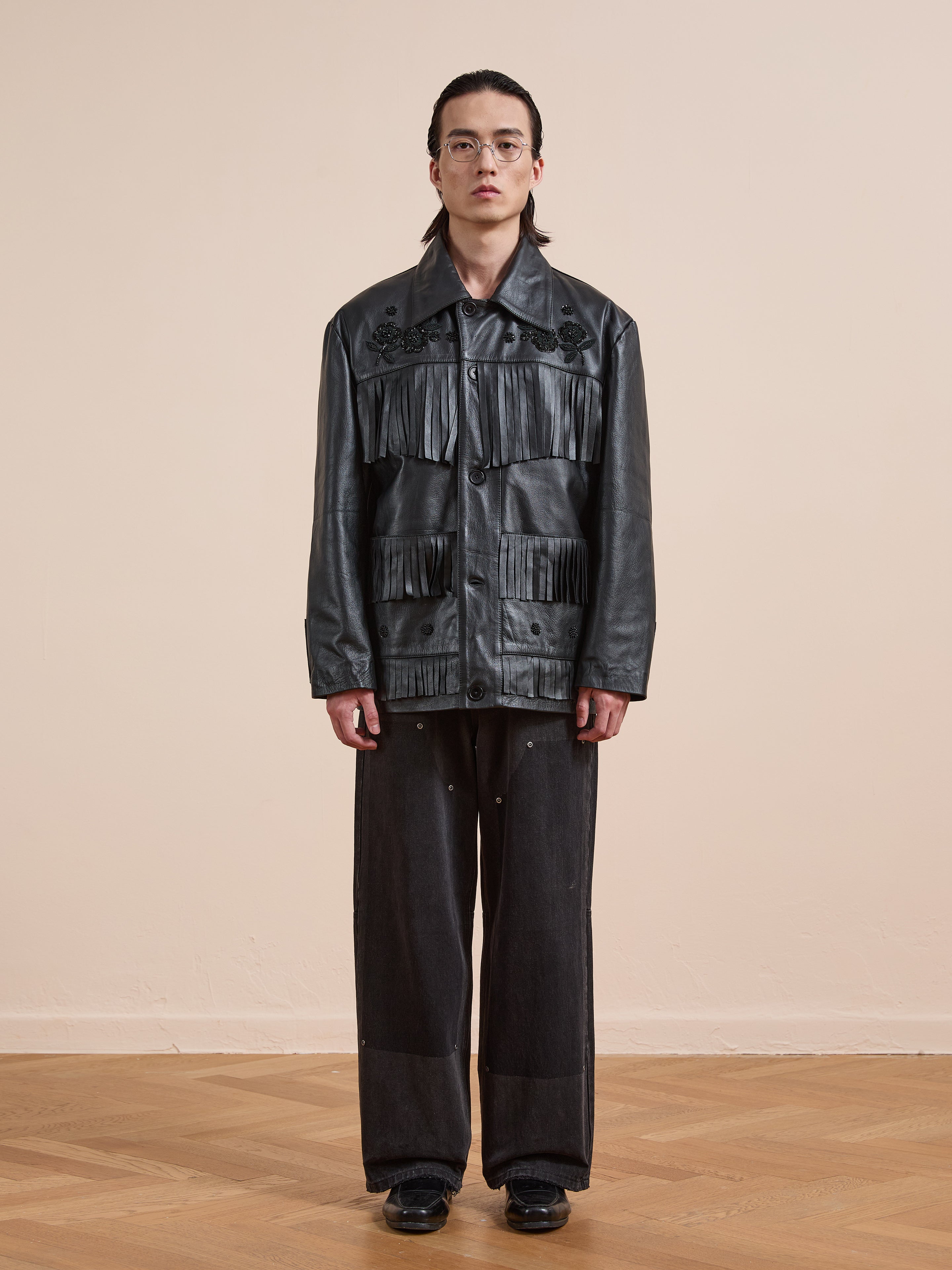 A person stands indoors against a plain background, wearing the FOUND Hand Embroidered Leather Fringe Jacket with fringe details, styled with black wide-leg pants.