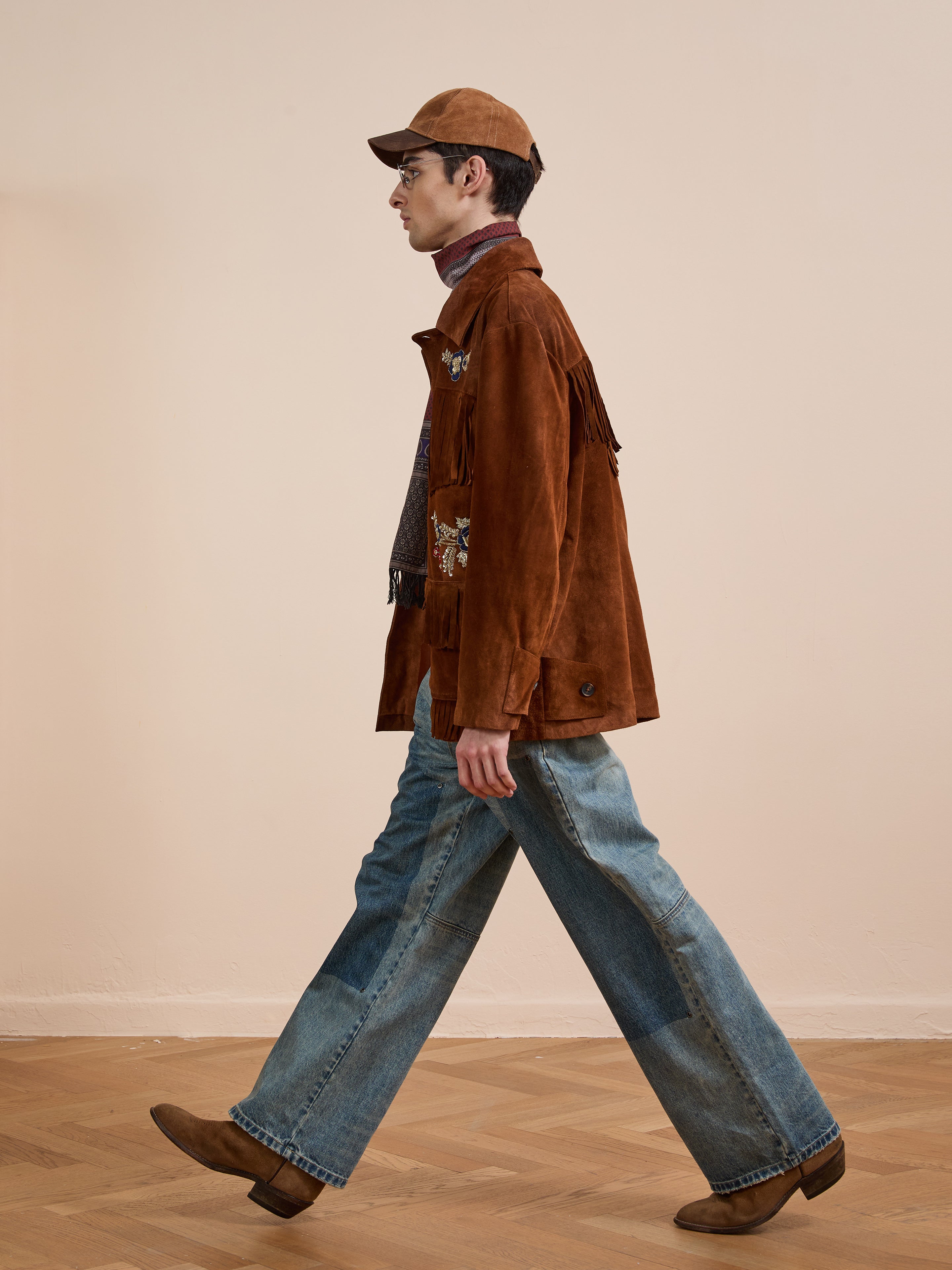 A person in the FOUND Hand Embroidered Suede Fringe Jacket, blue jeans, brown boots, and a cap walks across a wooden floor with a plain beige background.