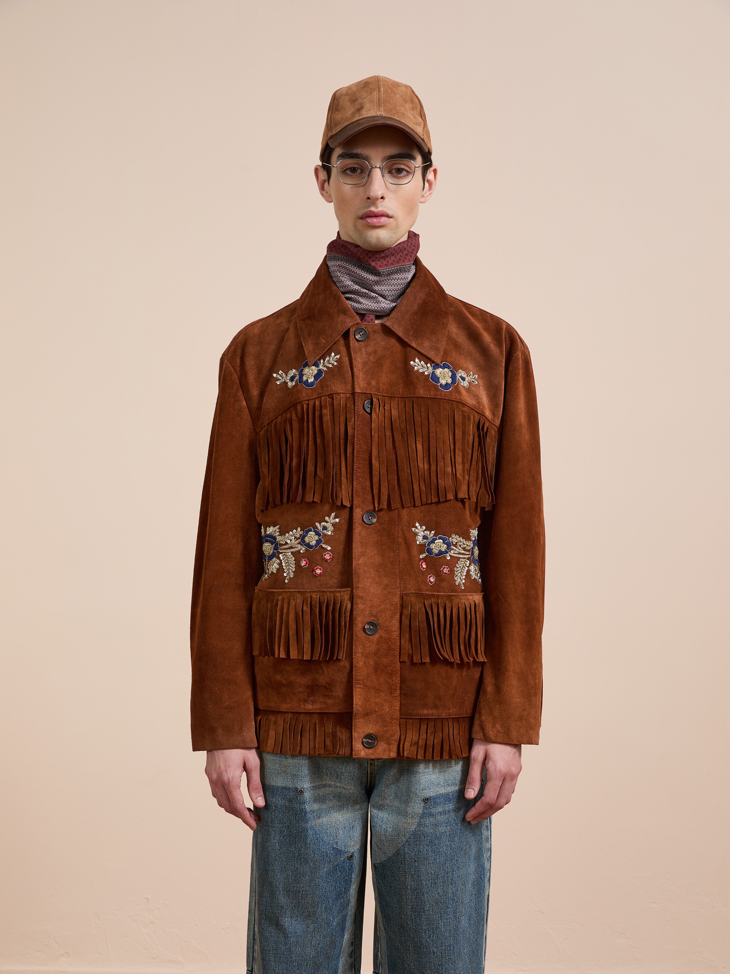 A person in glasses and a brown FOUND Hand Embroidered Suede Fringe Jacket, blue jeans, turtleneck, and brown cap stands before a plain beige background.