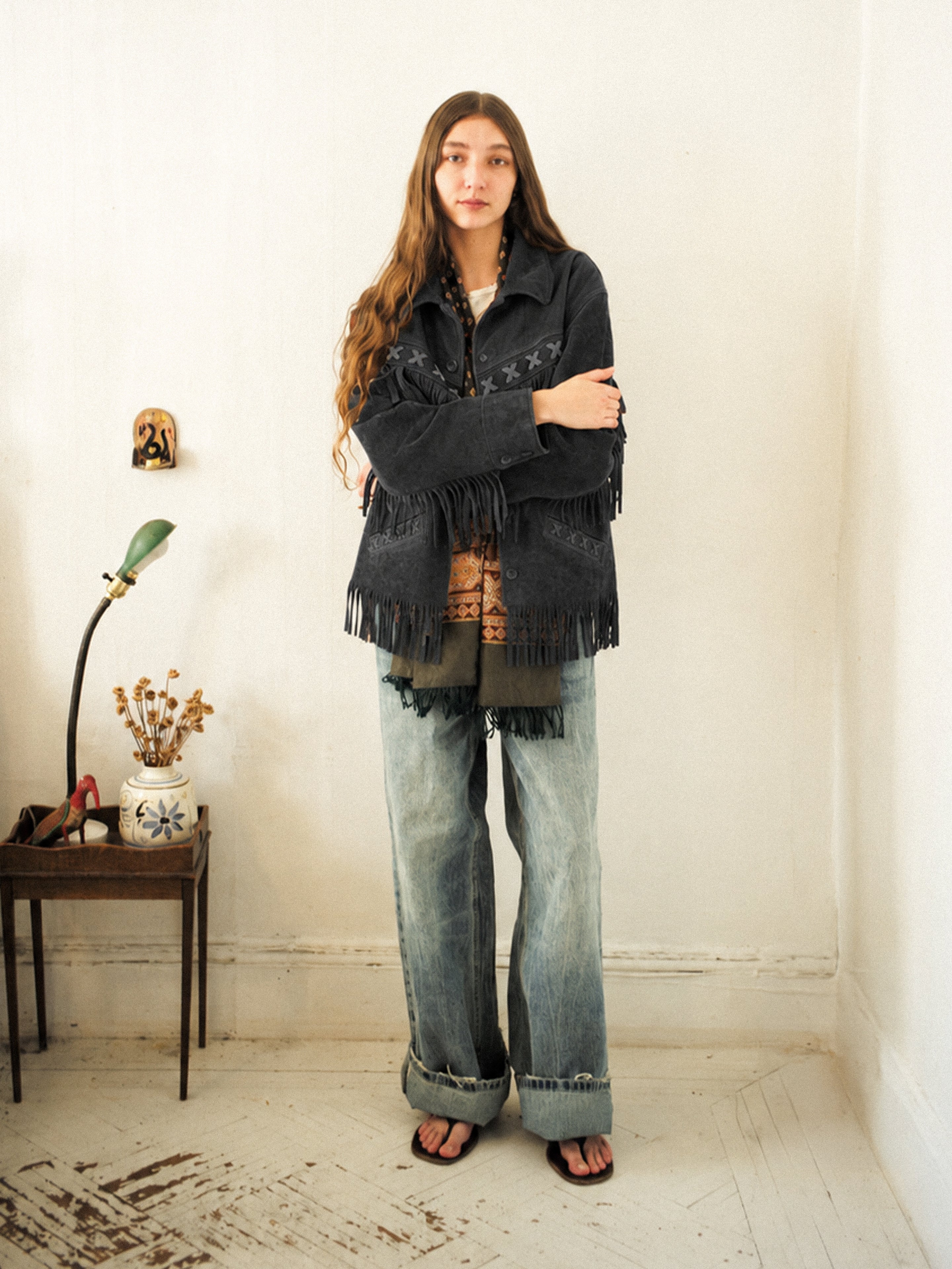 A woman with long hair stands indoors wearing the FOUND Two-Tone Western Fringe Suede Leather Jacket, paired with loose cuffed jeans, flip-flops, and layered tops. Nearby sits a small table topped with a lamp and dried flowers.