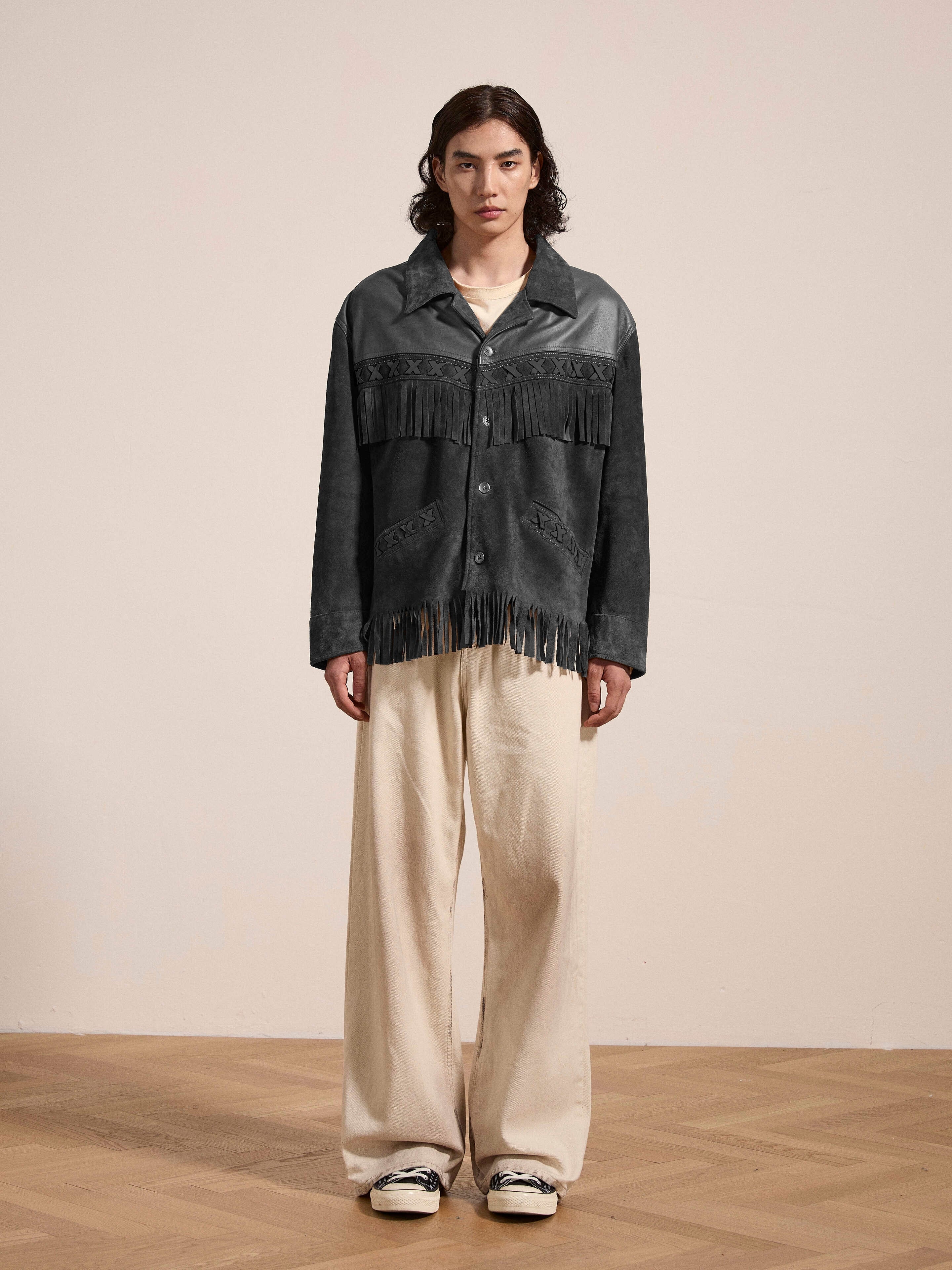 A person stands indoors on a wooden floor against a plain light-colored wall, wearing the FOUND Two-Tone Western Fringe Suede Leather Jacket paired with cream wide-leg pants and sneakers.