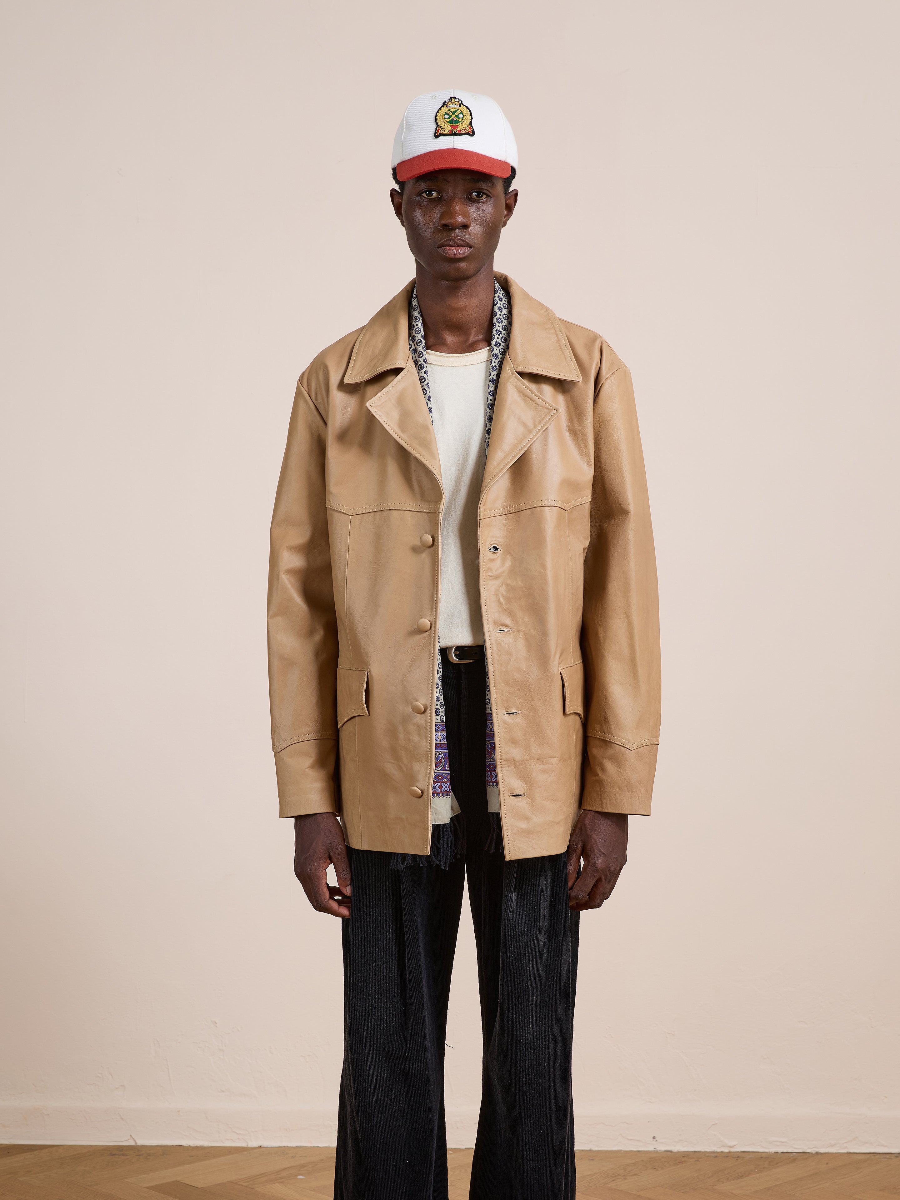 A person stands against a plain wall wearing the FOUND Western Oversized Leather Blazer in tan, paired with a white shirt, dark wide-leg pants, and a white baseball cap with a colorful logo.