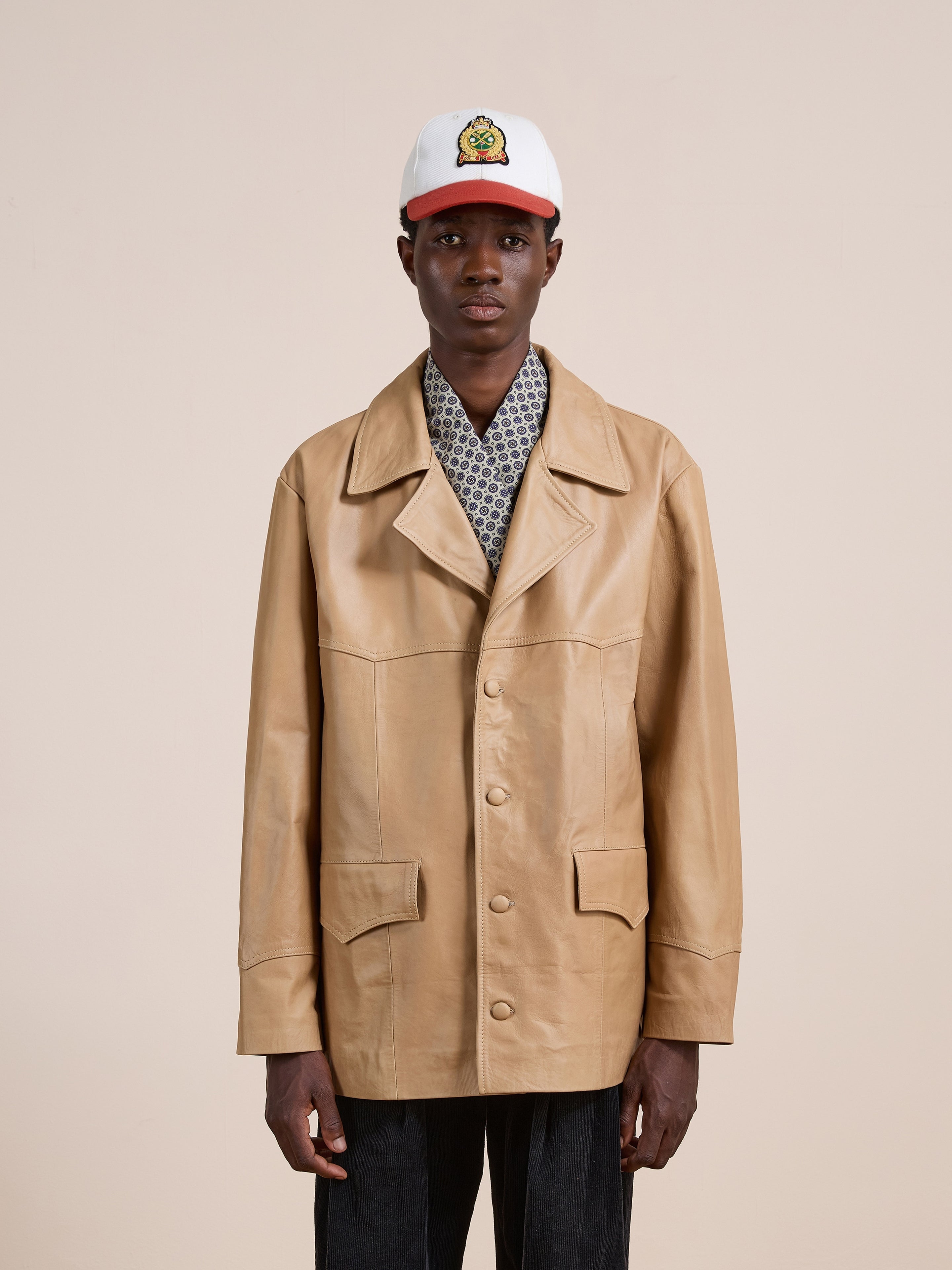 A person stands facing forward, wearing the FOUND Western Oversized Leather Blazer in tan over a patterned shirt, paired with dark pants and a white and red baseball cap featuring a yellow cartoon graphic on the front.