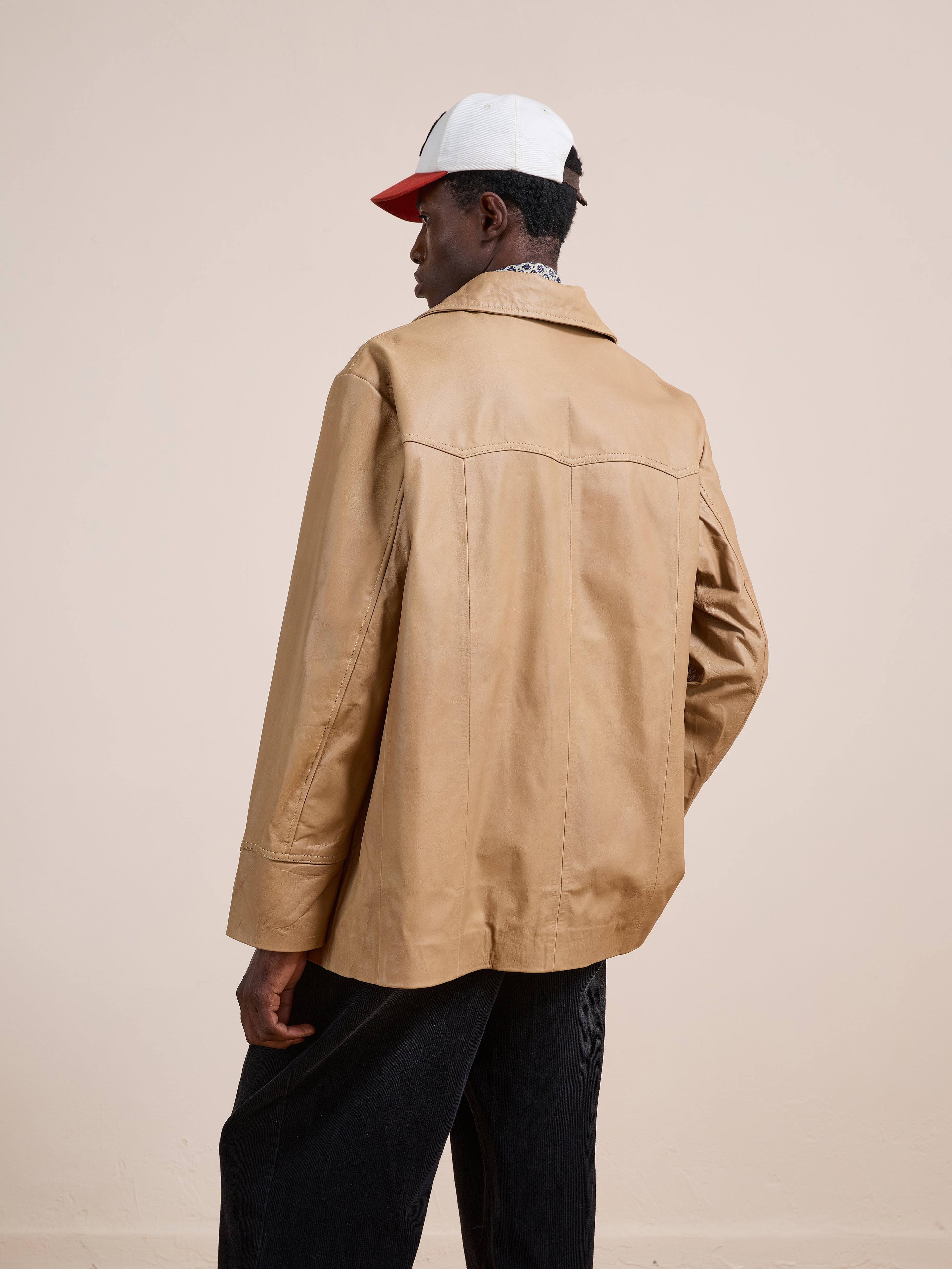 A person wearing the FOUND Western Oversized Leather Blazer stands with their back to a plain beige background, paired with black pants and a white and red baseball cap.