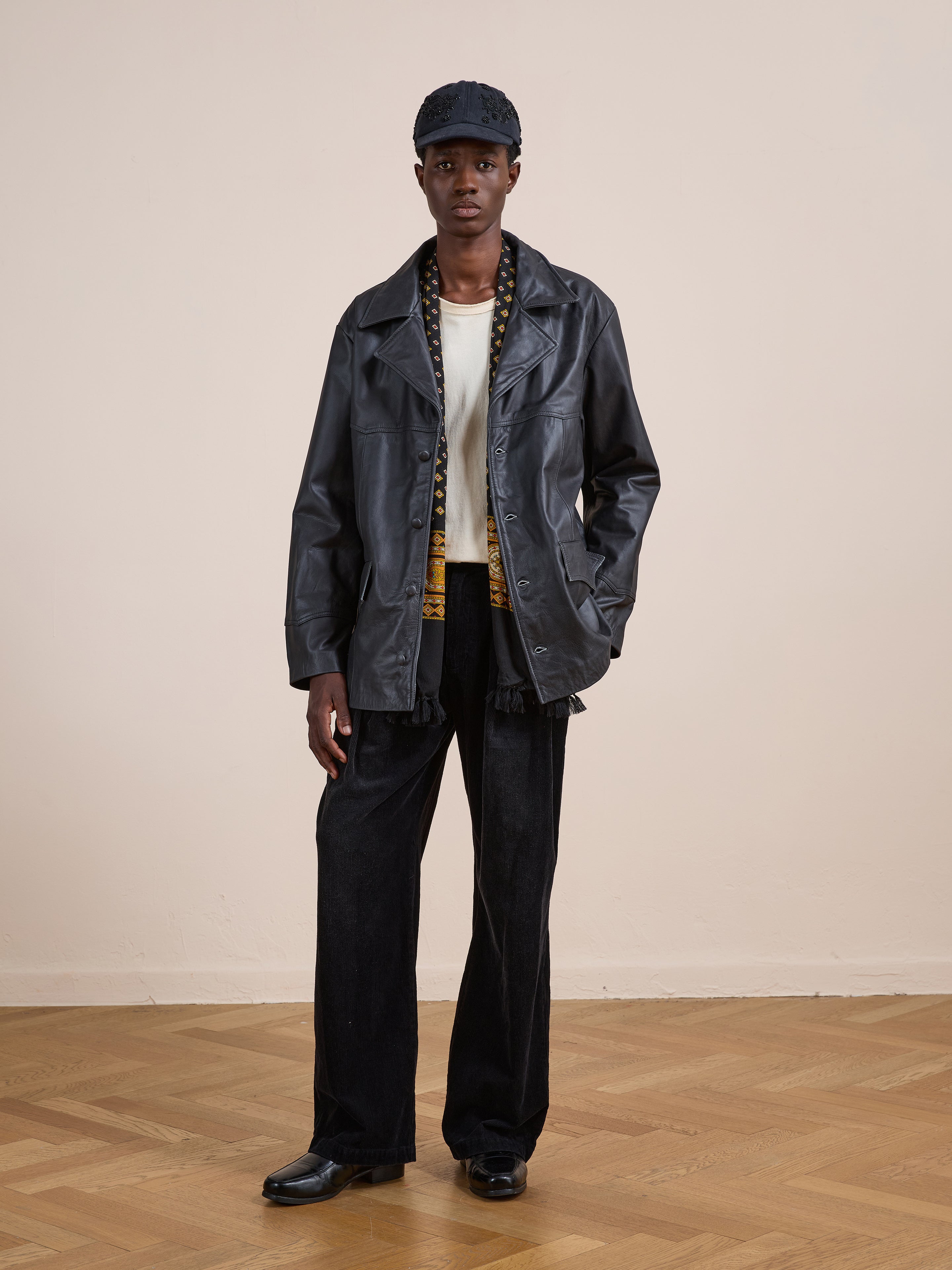 Dressed in FOUND's Western Oversized Leather Blazer, a person pairs it with a white shirt, patterned vest, black pants, black shoes, and a black cap while standing on a wooden floor against a plain beige wall.