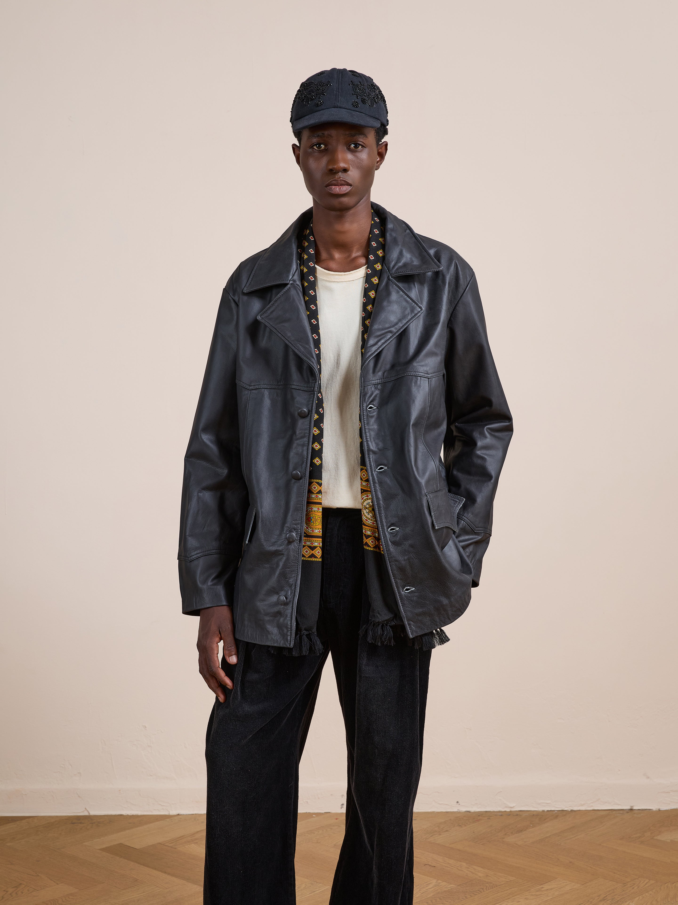 A person stands indoors with hands in their pockets, wearing FOUND’s Western Oversized Leather Blazer over a white shirt, dark pants, and a black cap, set against a plain background.