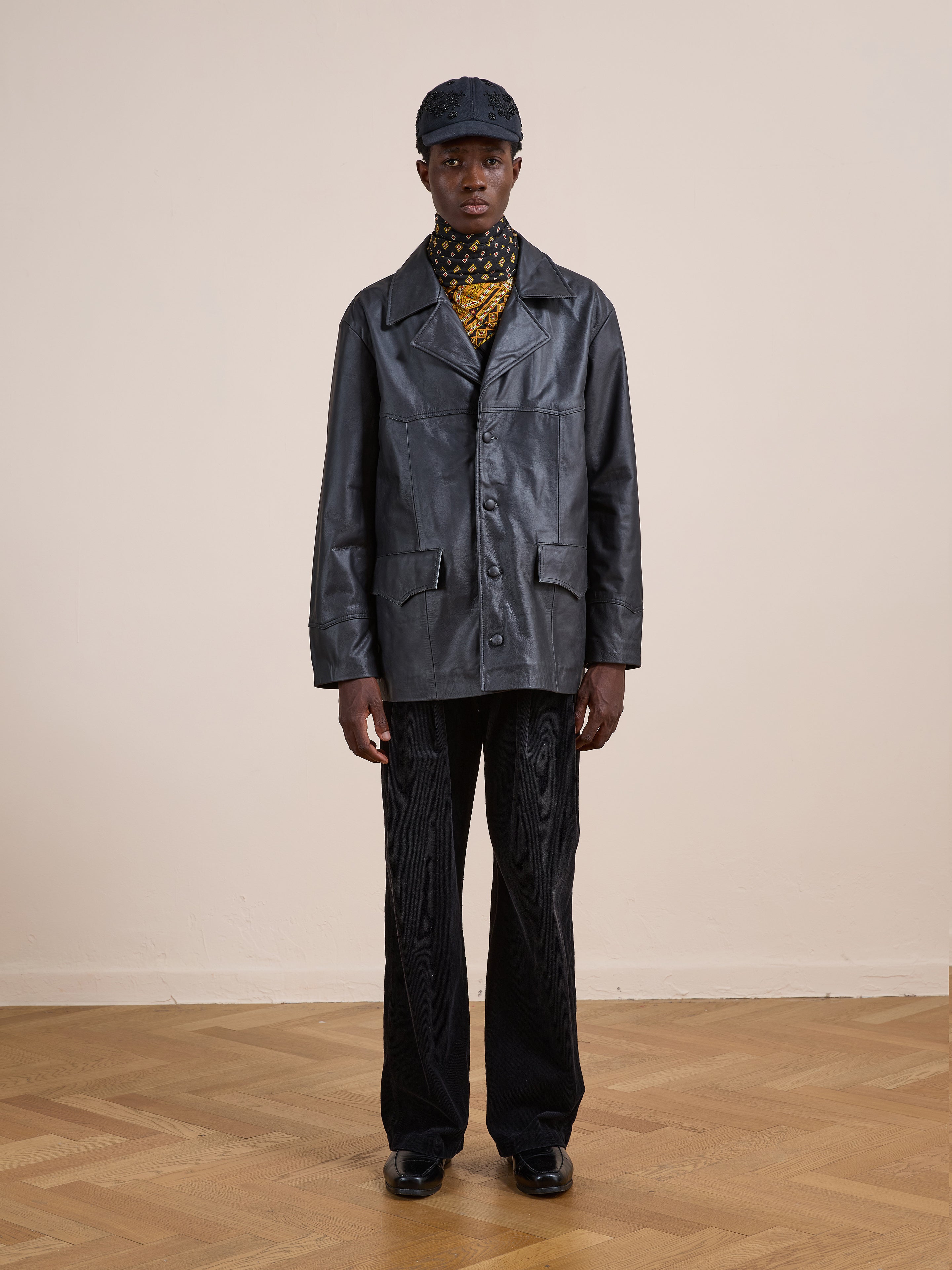 Indoors on a parquet floor and plain beige wall, a person wears the FOUND Western Oversized Leather Blazer, styled with a patterned turtleneck, black pants, black shoes, and a dark cap.