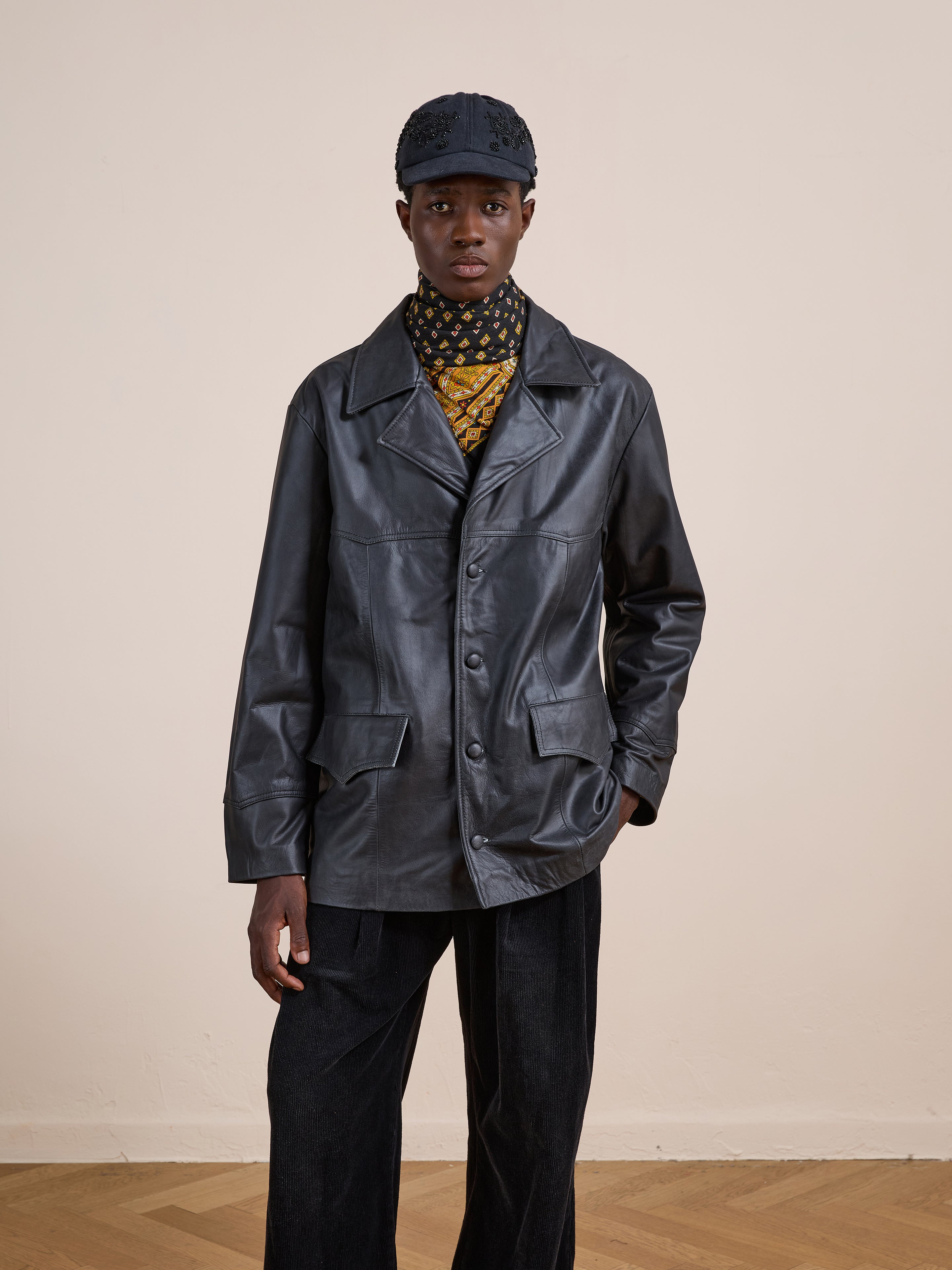Person wearing FOUND's Western Oversized Leather Blazer, paired with a patterned scarf, black cap, and black pants, standing indoors against a plain beige wall.