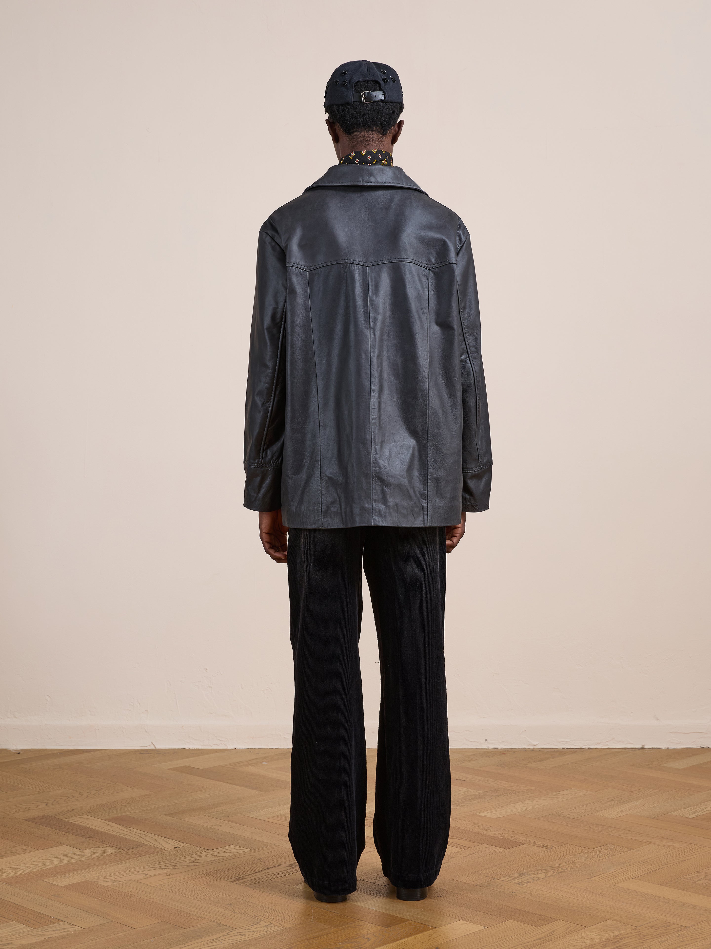 A person stands facing away, wearing the FOUND Western Oversized Leather Blazer with dark wide-leg pants and a cap, on a light wooden floor against a plain light-colored wall.