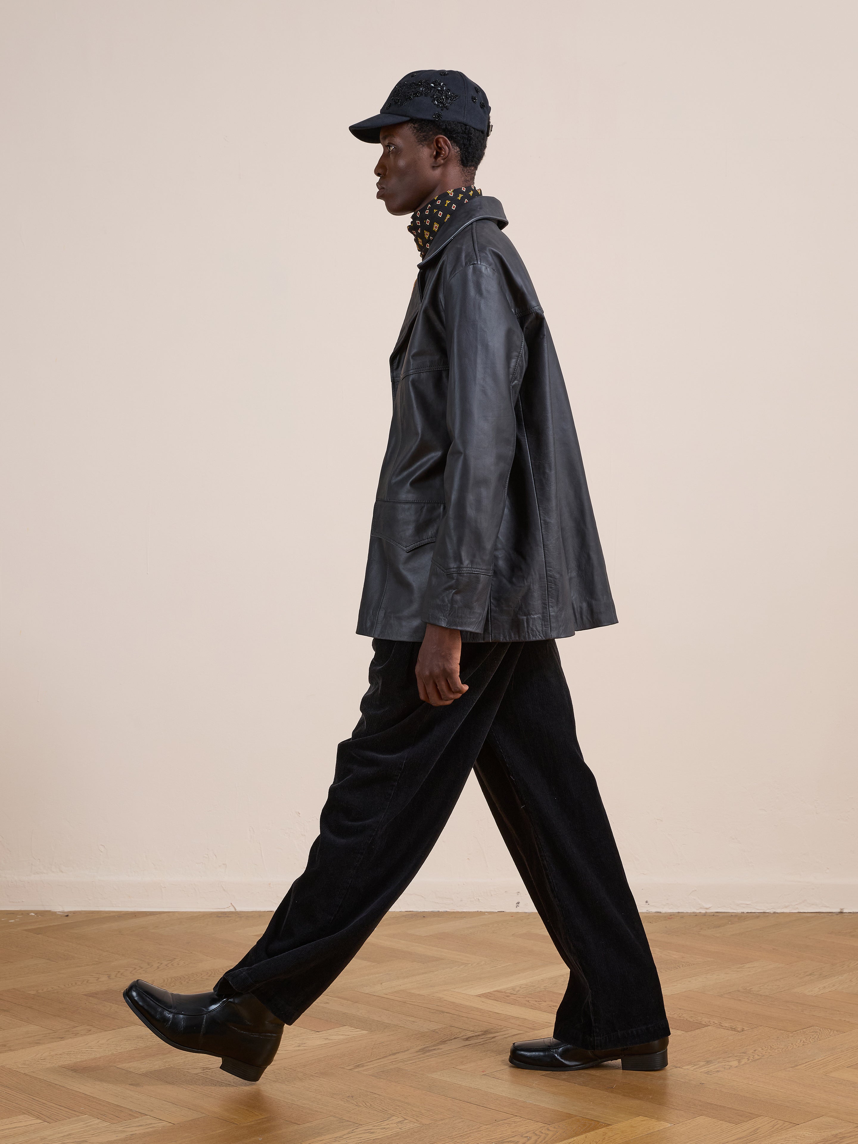 A person wearing the FOUND Western Oversized Leather Blazer, black pants, black shoes, and a black cap walks indoors on a parquet floor against a plain light-colored wall.