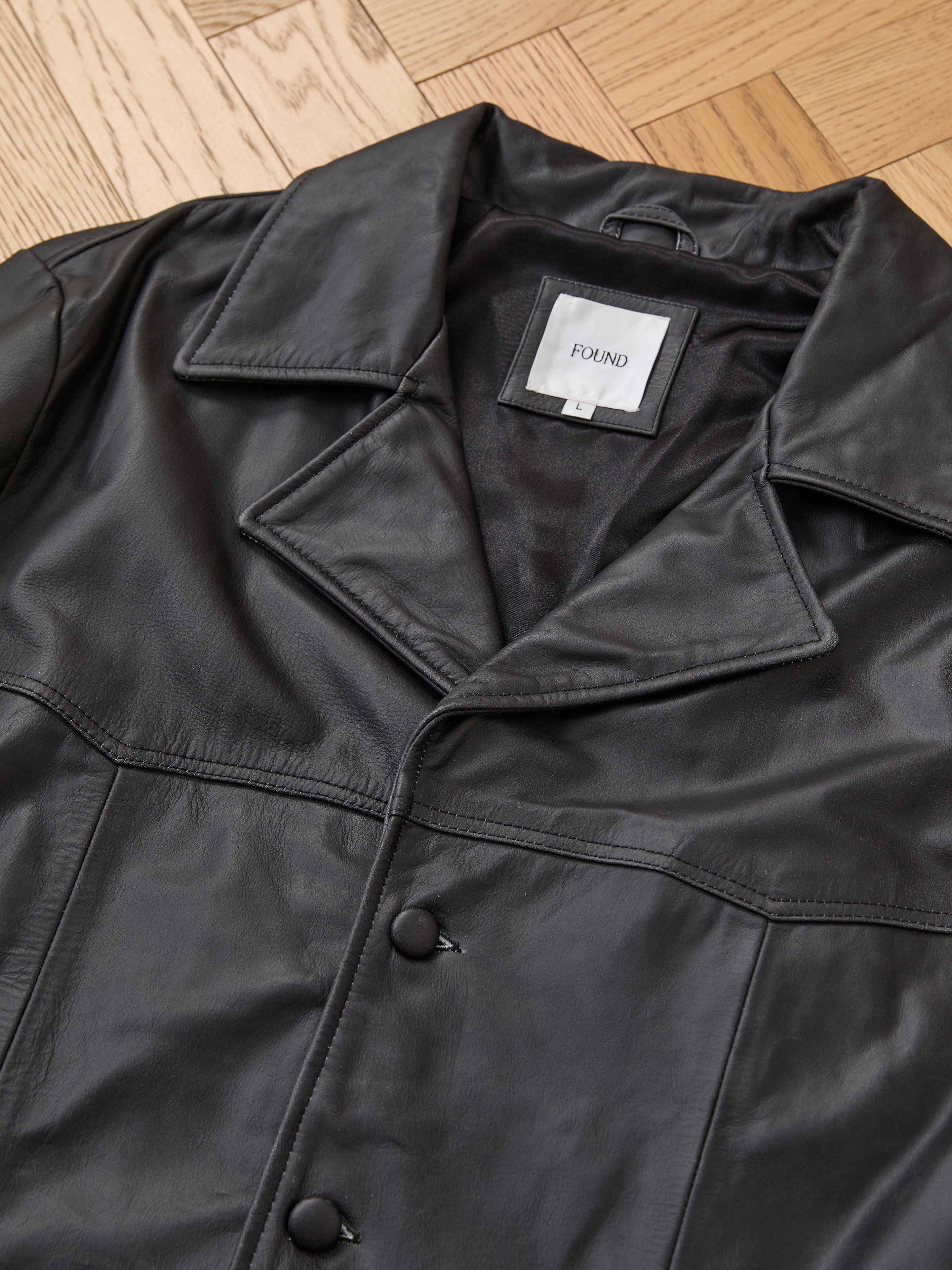 The FOUND Western Oversized Leather Blazer in black vintage leather features a wide collar and button closures, shown laid on a wooden floor with an inside white FOUND label visible.