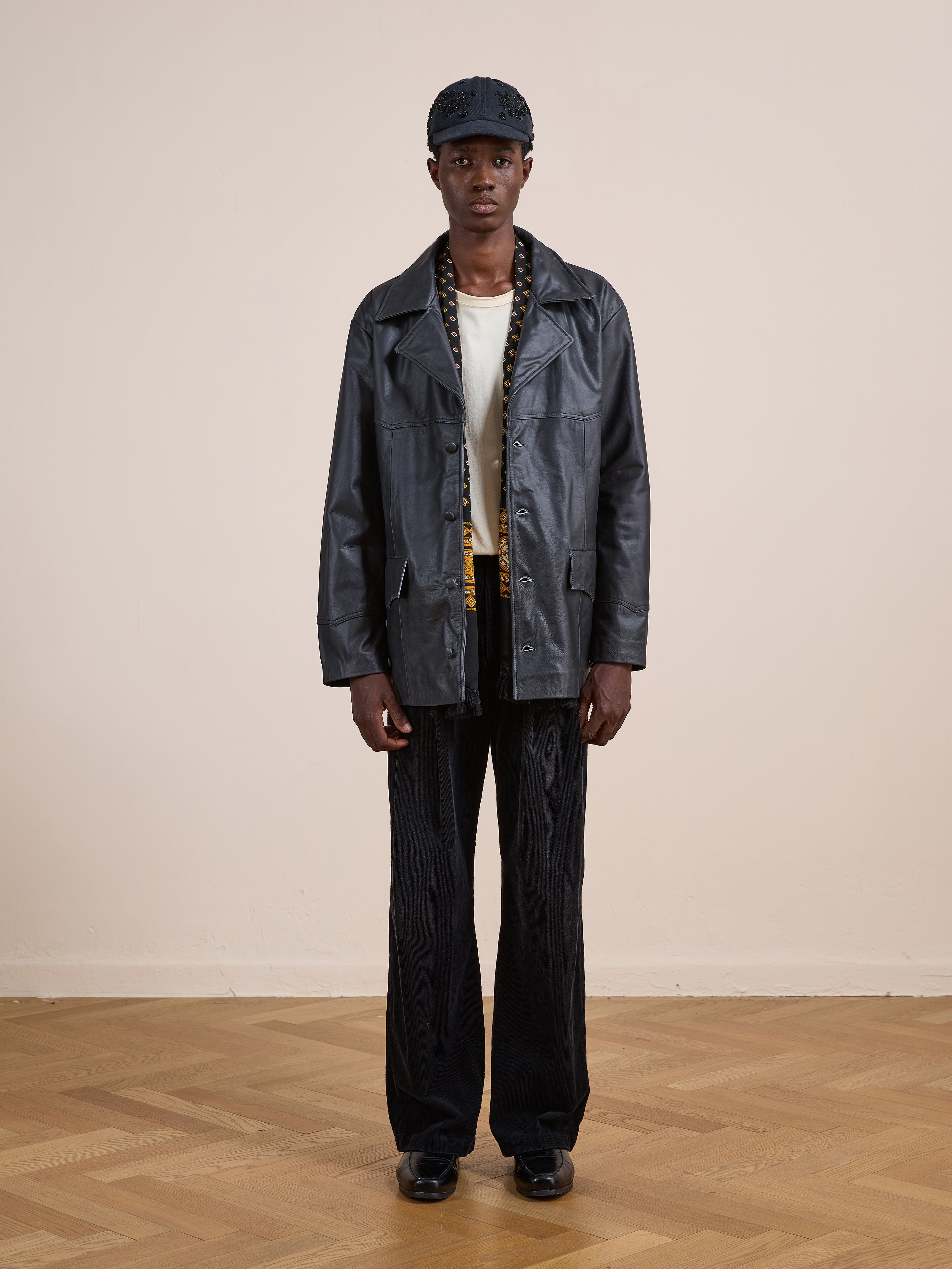 A person stands indoors on a wooden floor, facing the camera against a beige wall, wearing the FOUND Western Oversized Leather Blazer with black pants, a light shirt, black cap, and black shoes.
