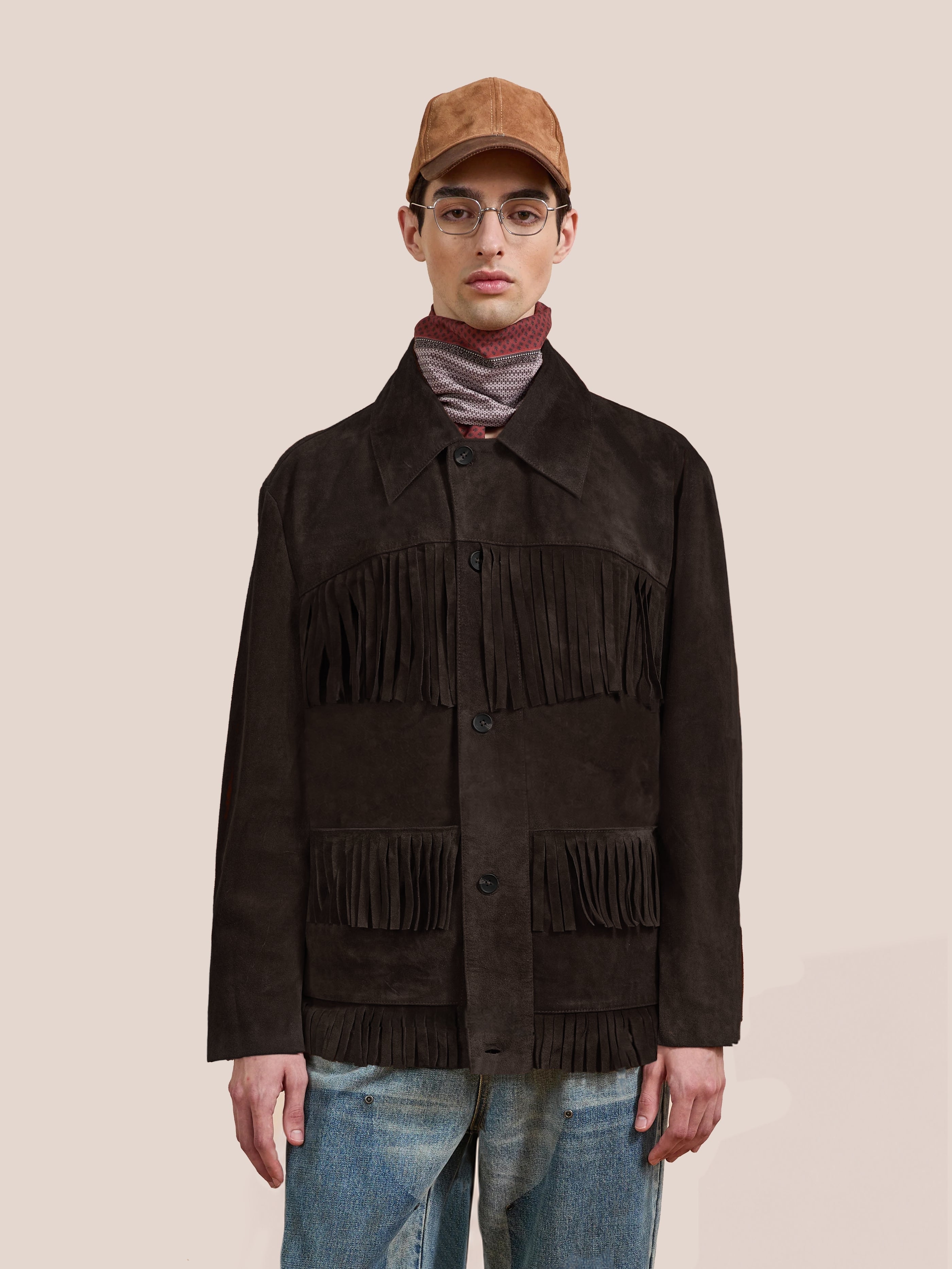 A person wearing the FOUND Western Suede Fringe Jacket, along with a striped scarf, light brown cap, glasses, and blue jeans, stands against a plain beige background.