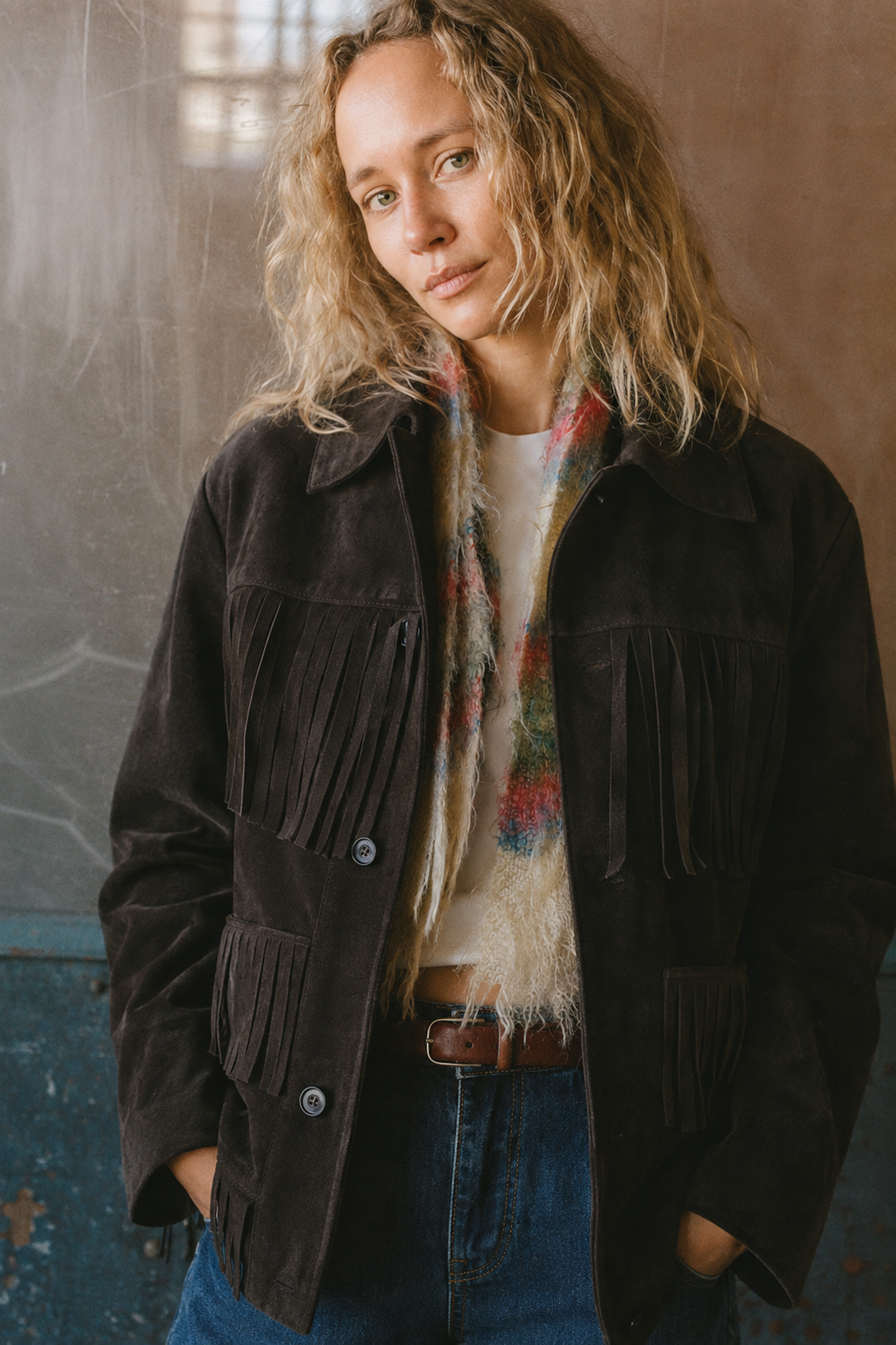 A person with wavy blonde hair wears the FOUND Western Suede Fringe Jacket in dark brown, styled with a multicolored scarf and blue jeans while standing against a textured wall.
