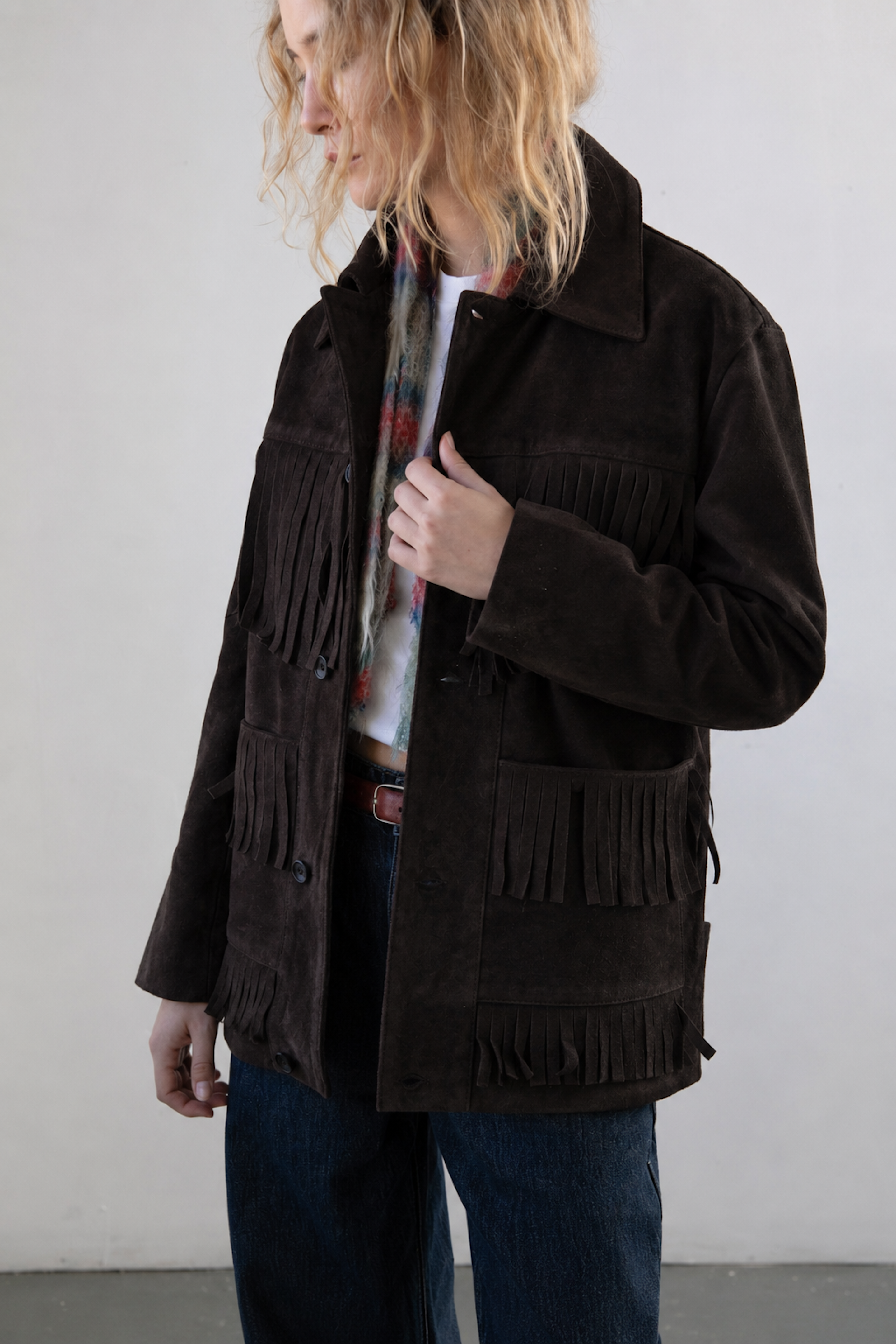 A person with wavy blond hair wears the FOUND Western Suede Fringe Jacket in dark brown over a patterned shirt and blue jeans, standing against a plain light background.