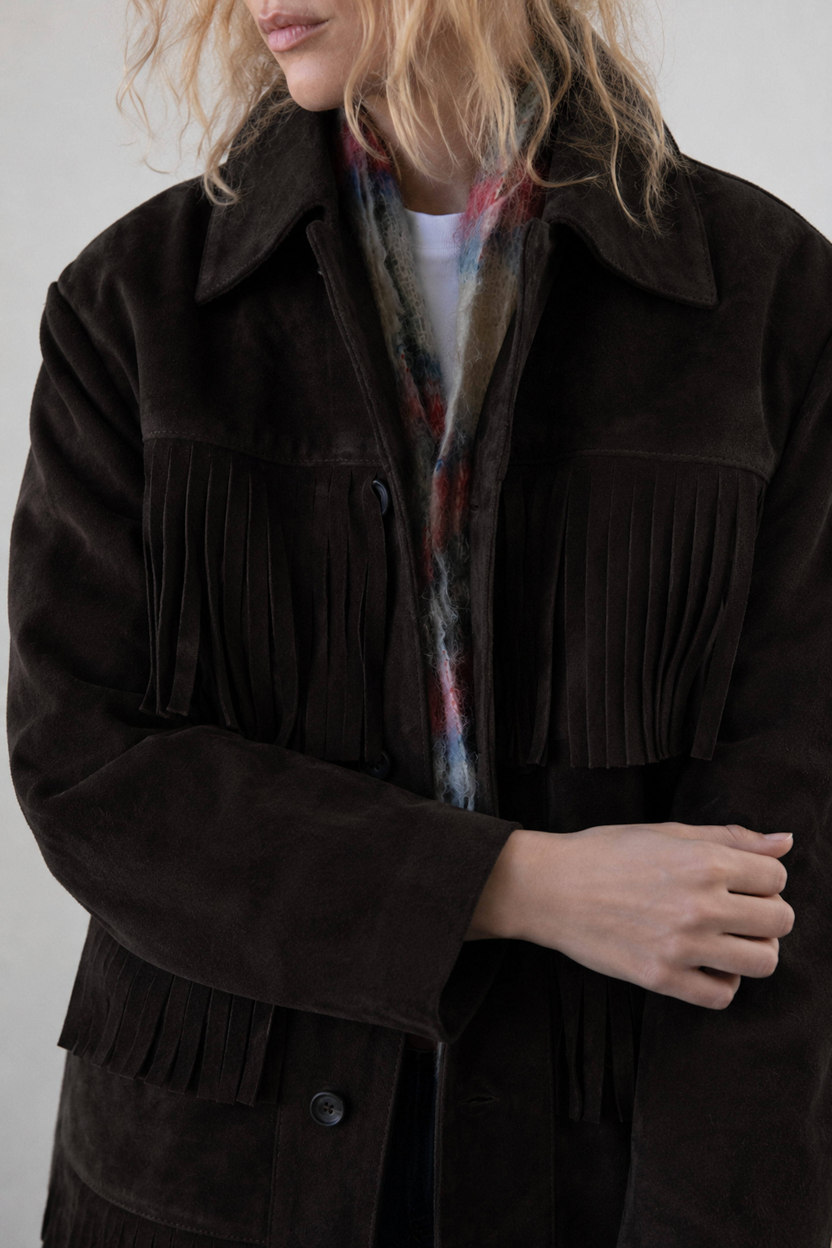 A woman with wavy blonde hair wears the FOUND Western Suede Fringe Jacket in dark brown over a white shirt and a multicolored scarf. Her face is partially out of frame.