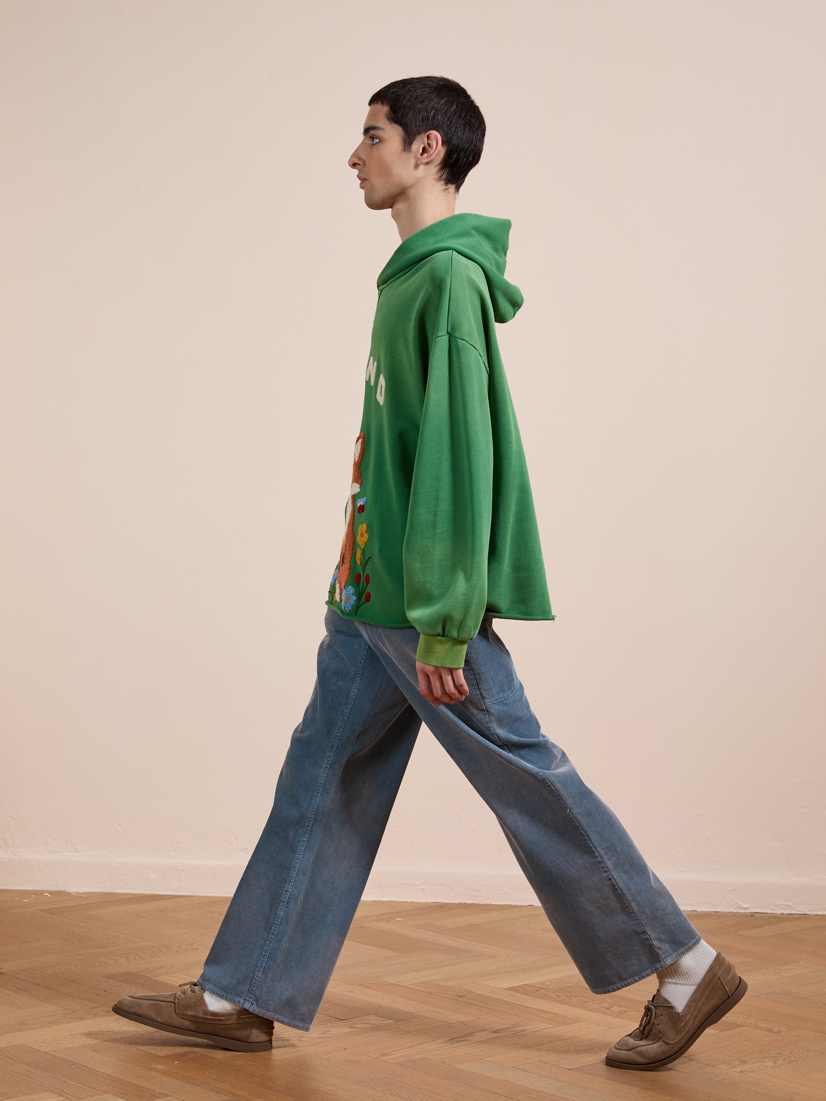 A young person with short dark hair walks indoors on a wooden floor with a plain beige background, wearing a green FOUND Embroidered Wildlife Hoodie, wide-leg blue jeans, white socks, and brown shoes.