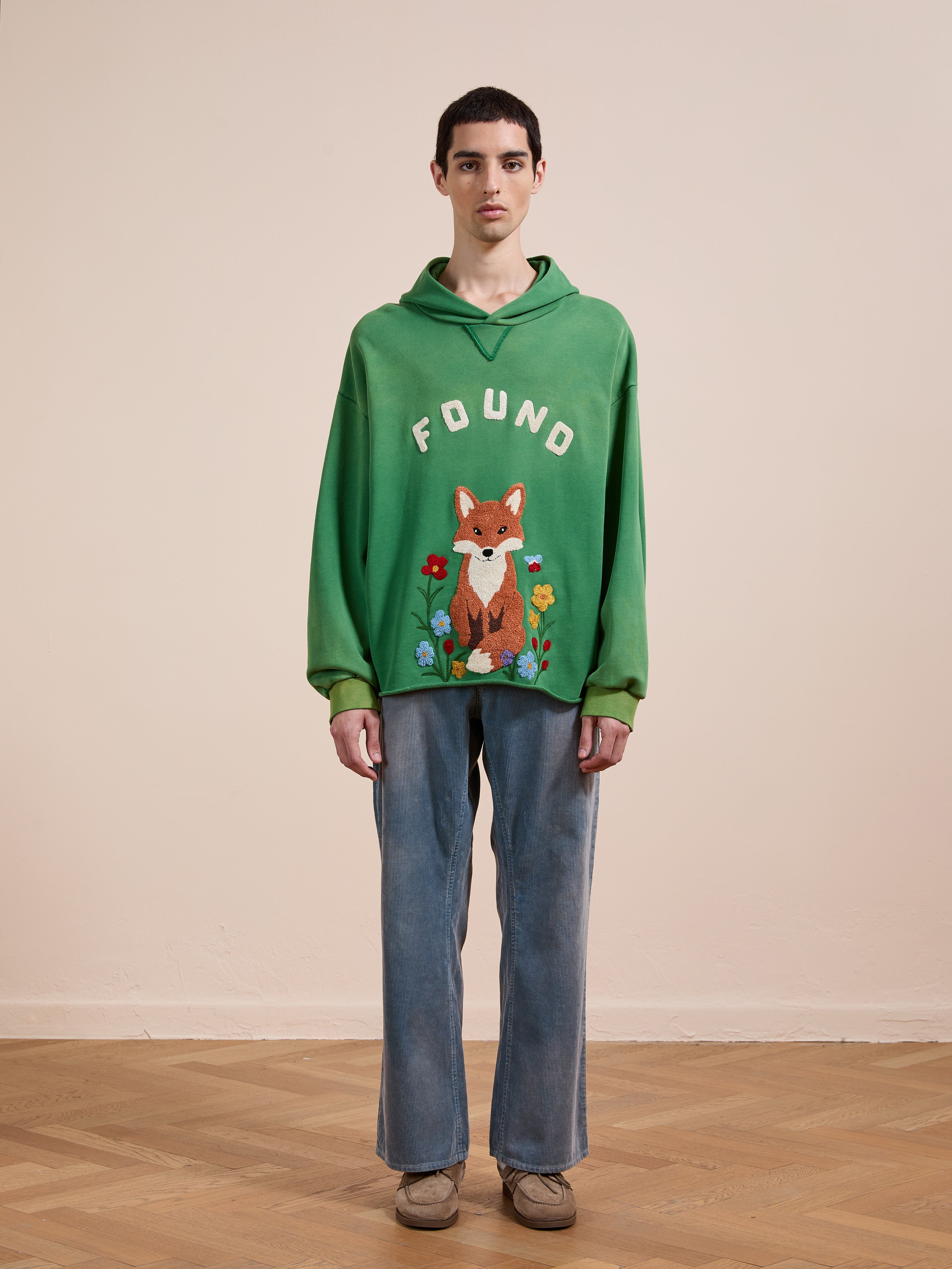 A person stands indoors against a plain background, wearing the FOUND Embroidered Wildlife Hoodie with a fox graphic, paired with blue jeans and brown shoes.