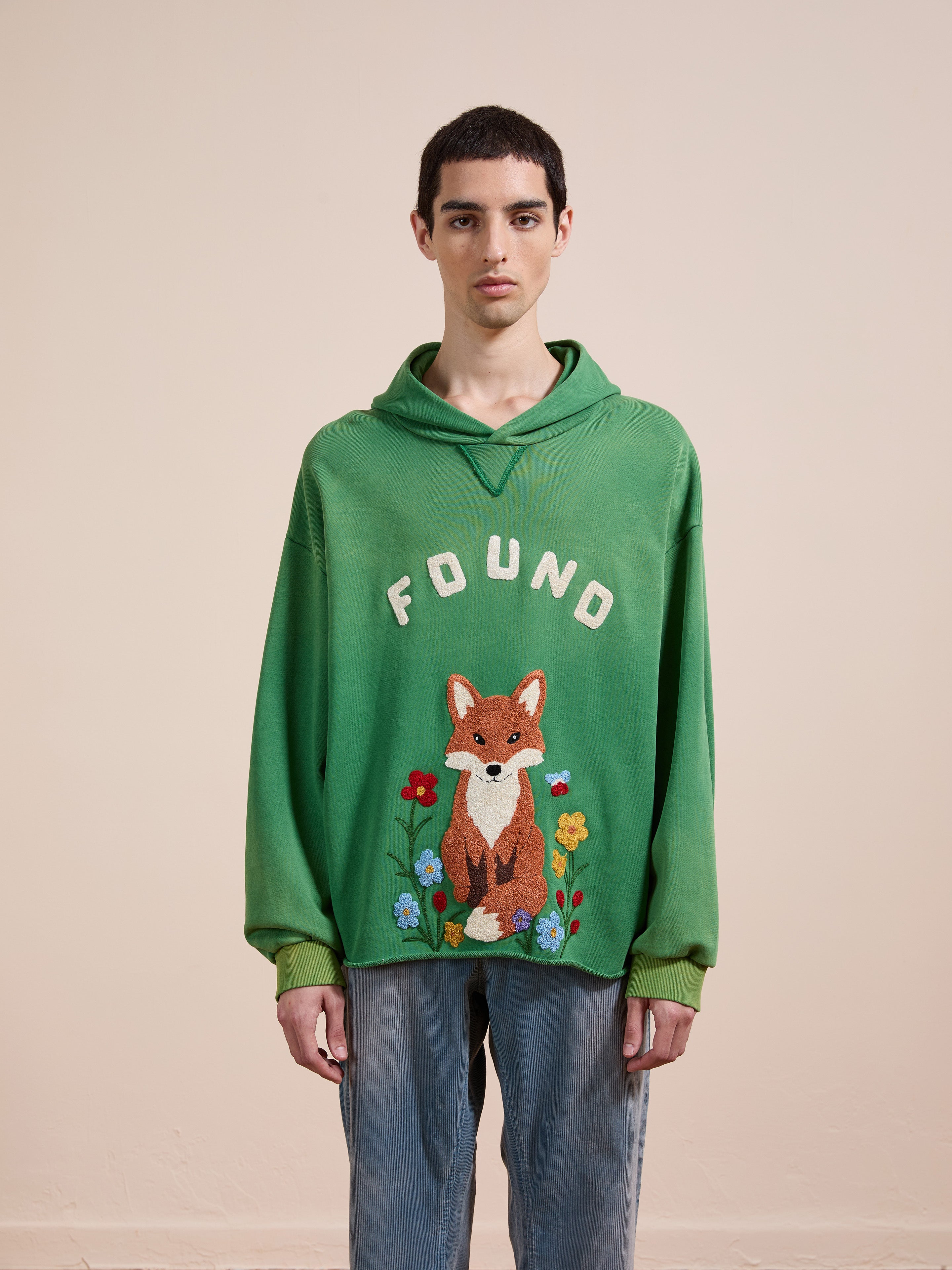 A person wears the FOUND Embroidered Wildlife Hoodie featuring an illustrated fox with flowers, paired with light blue jeans, standing against a plain beige background.