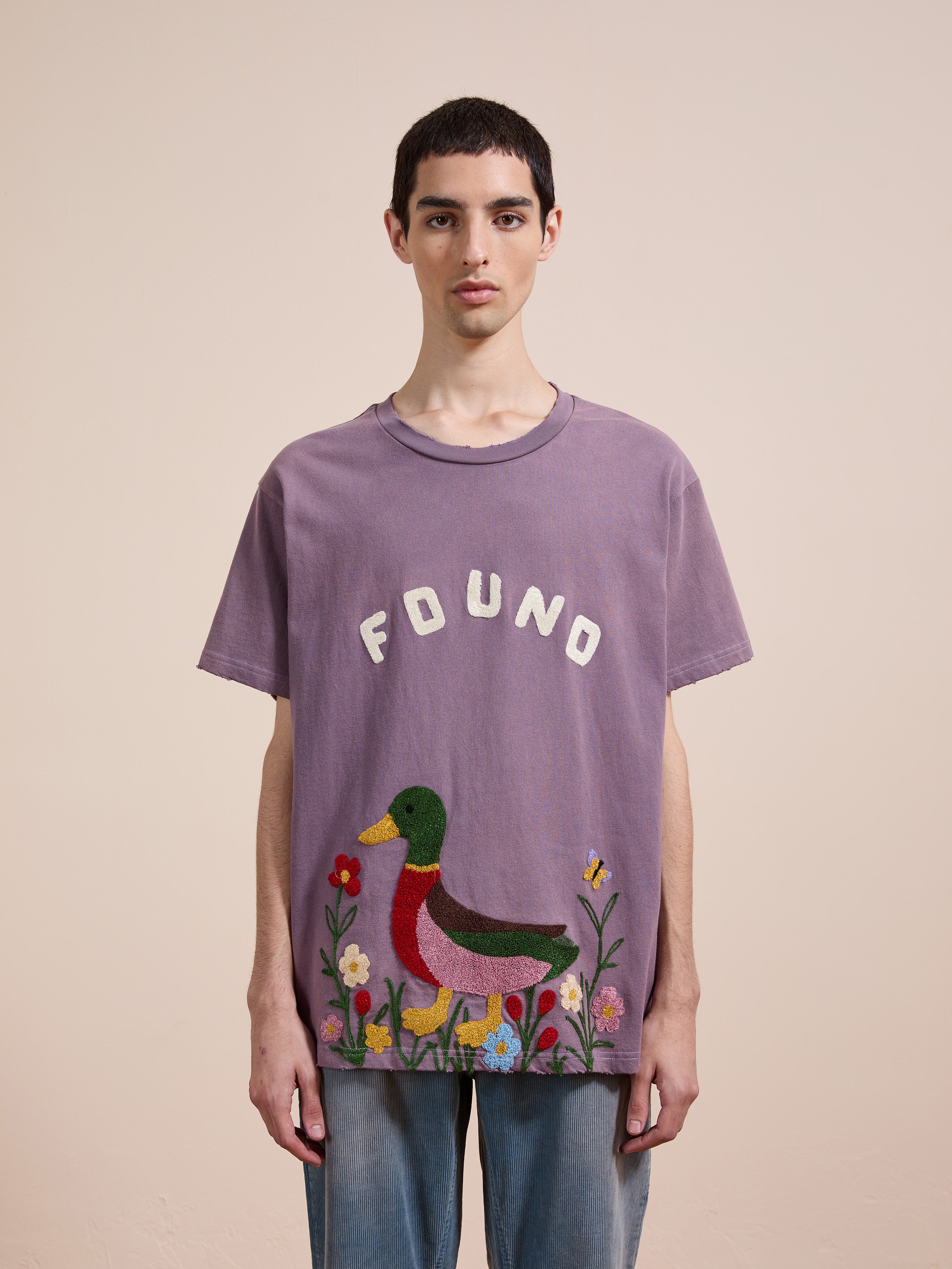 A person stands against a plain background wearing the FOUND Embroidered Wildlife Tee, which features a purple color and a vibrant duck with flowers graphic near the bottom.