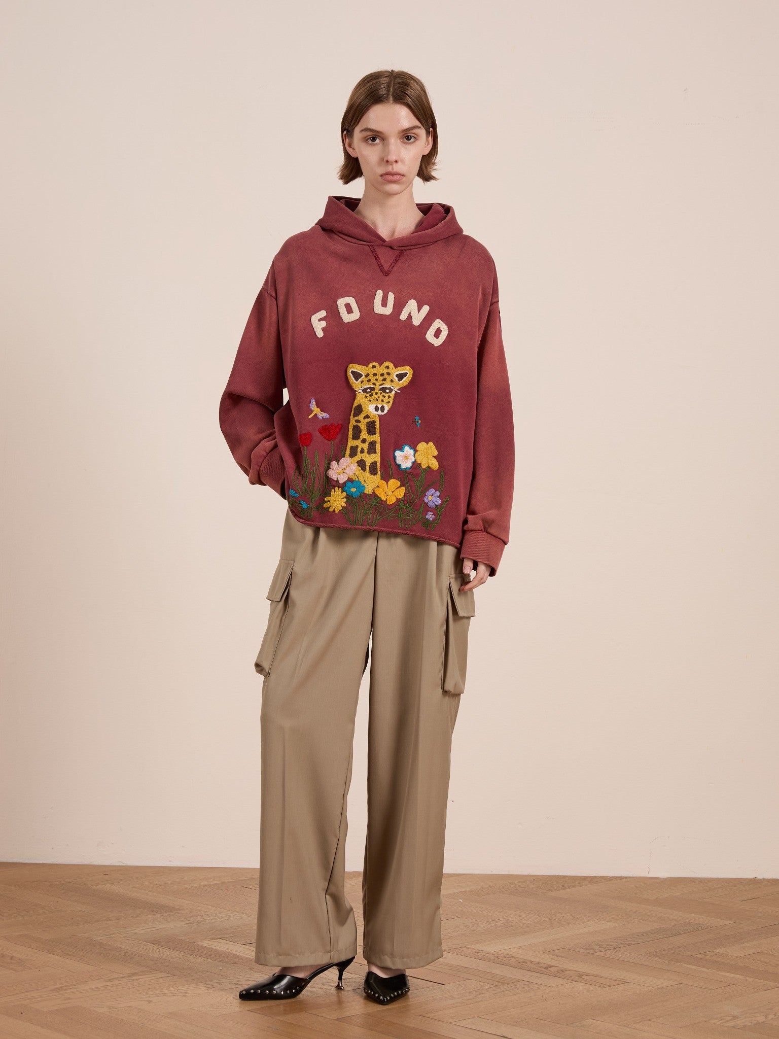 Indoors, someone wears the FOUND Embroidered Wildlife Hoodie in maroon with a giraffe graphic, styled with beige cargo pants and black heeled shoes.