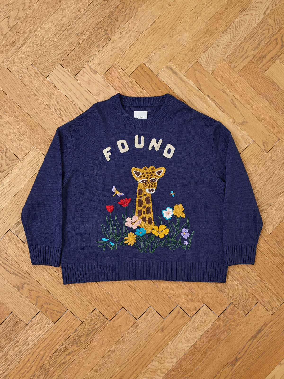 Wildlife Knit Sweater by FOUND featuring “FOUND” text, embroidered giraffe, flowers, and butterfly, displayed flat on a wooden herringbone floor.