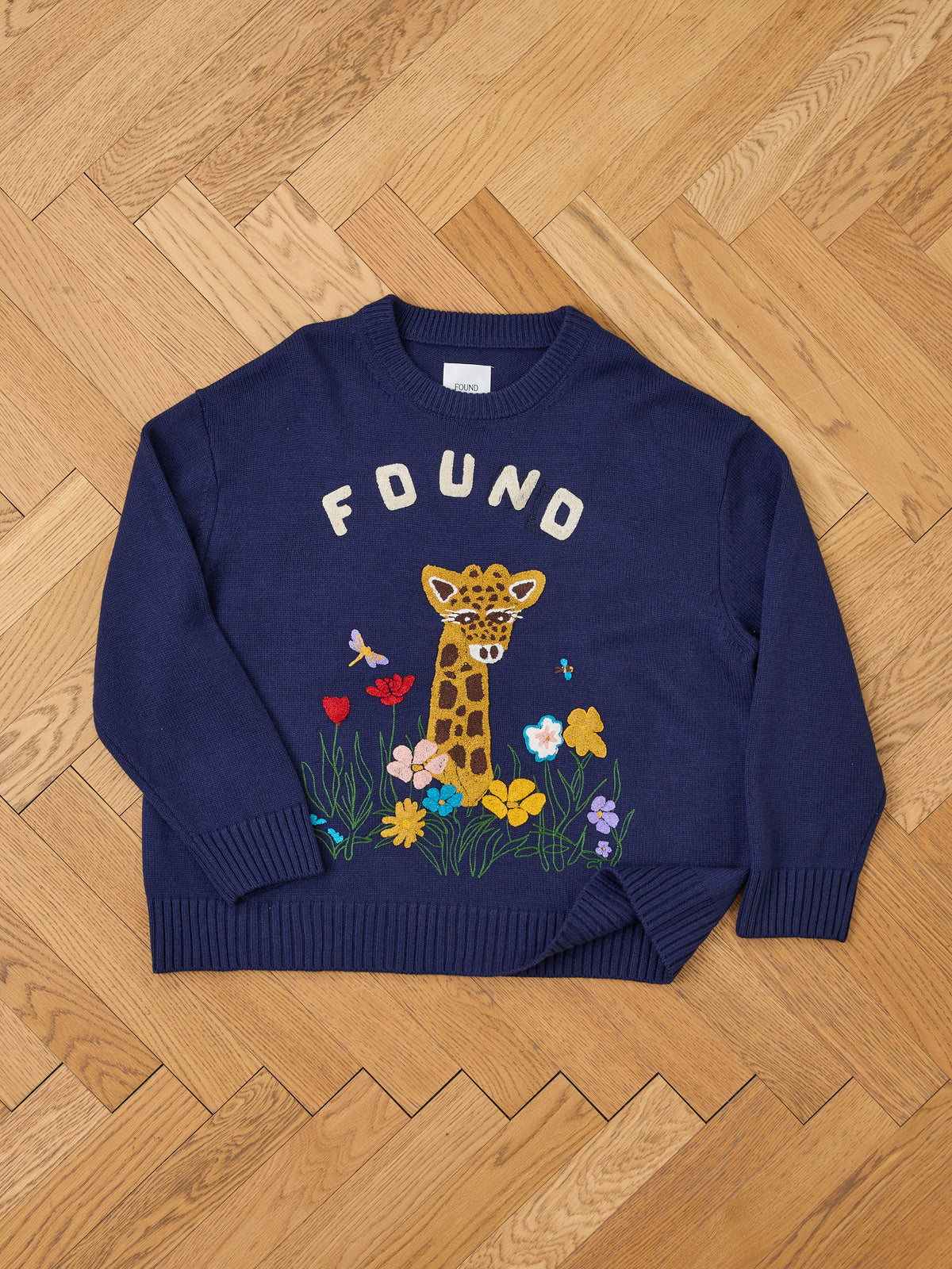 The Wildlife Knit Sweater by FOUND is a navy blue unisex crewneck with “FOUND,” a giraffe face, and colorful flowers embroidered on the front.