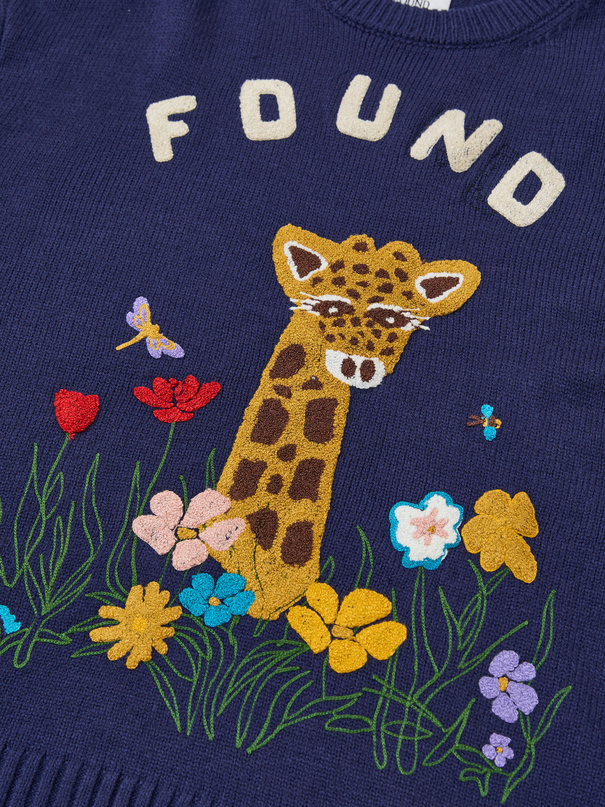 The Wildlife Knit Sweater by FOUND is a navy unisex crewneck featuring “FOUND,” an embroidered giraffe head, flowers, and small insects on the front.