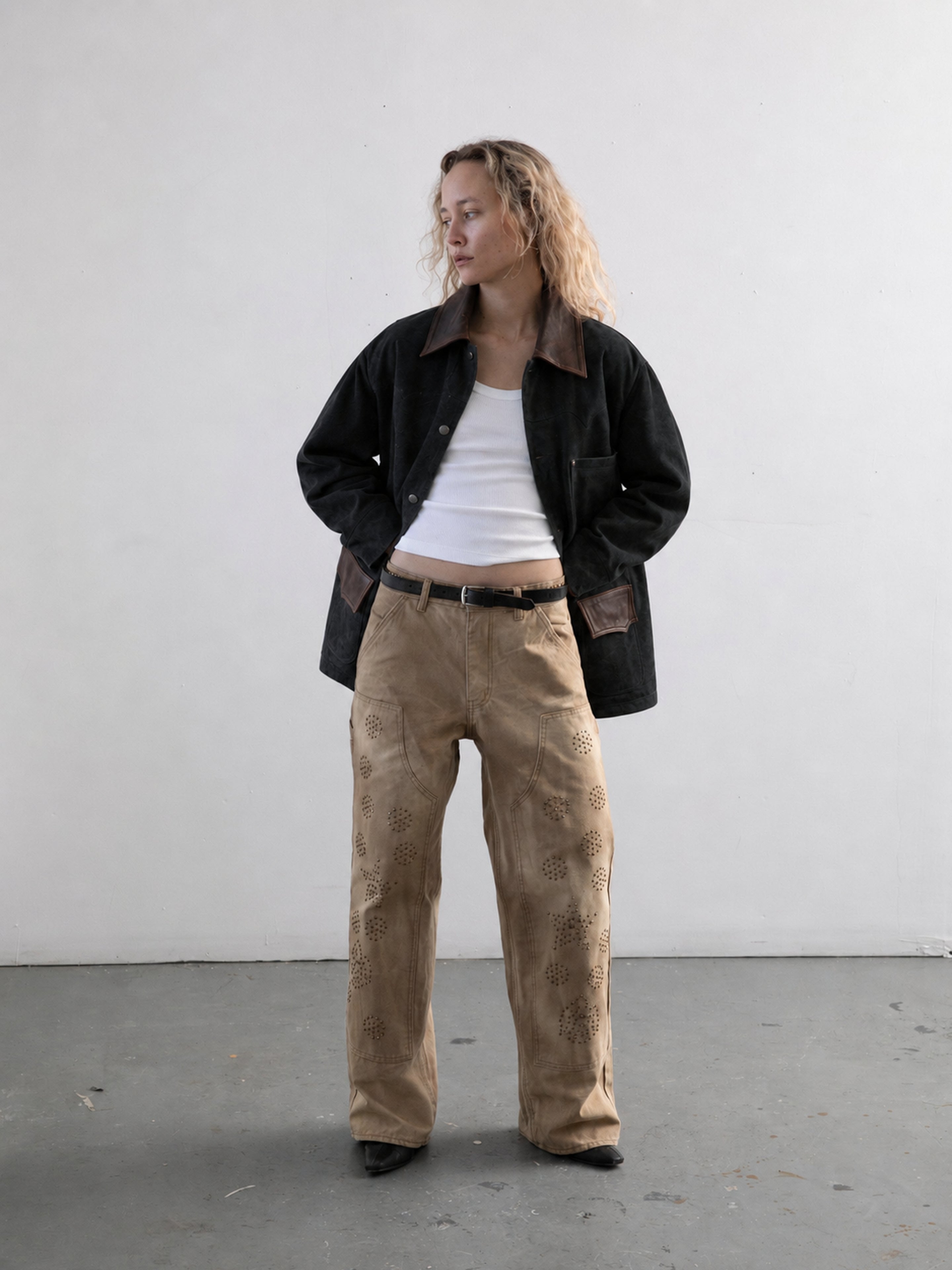 A person stands indoors against a plain background, looking to the side with hands in the pockets of the FOUND Suede Contrast Barn Jacket, worn over a white tank top and beige embroidered pants.