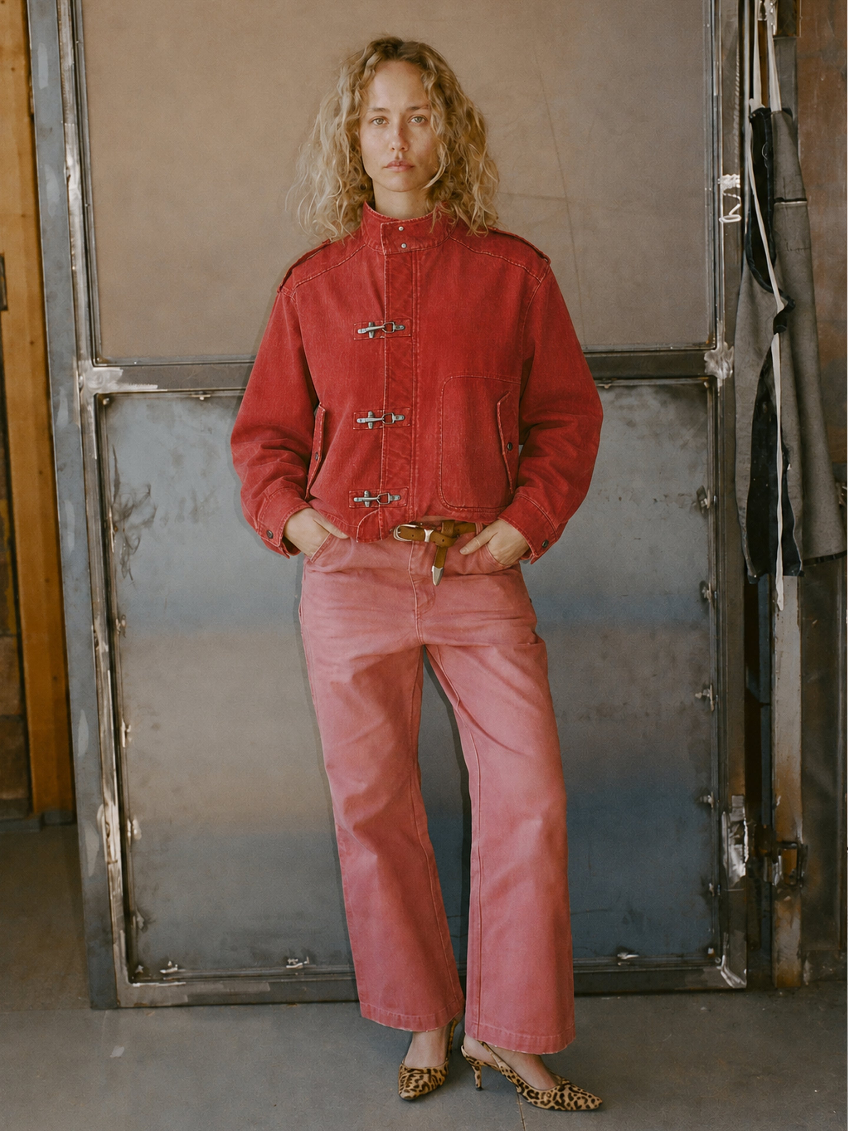 Person stands indoors wearing the FOUND Utility Clasp Work Jacket in vintage red with toggle closures, matching red pants, a brown belt, and leopard print heels, against a backdrop of metal and wood.