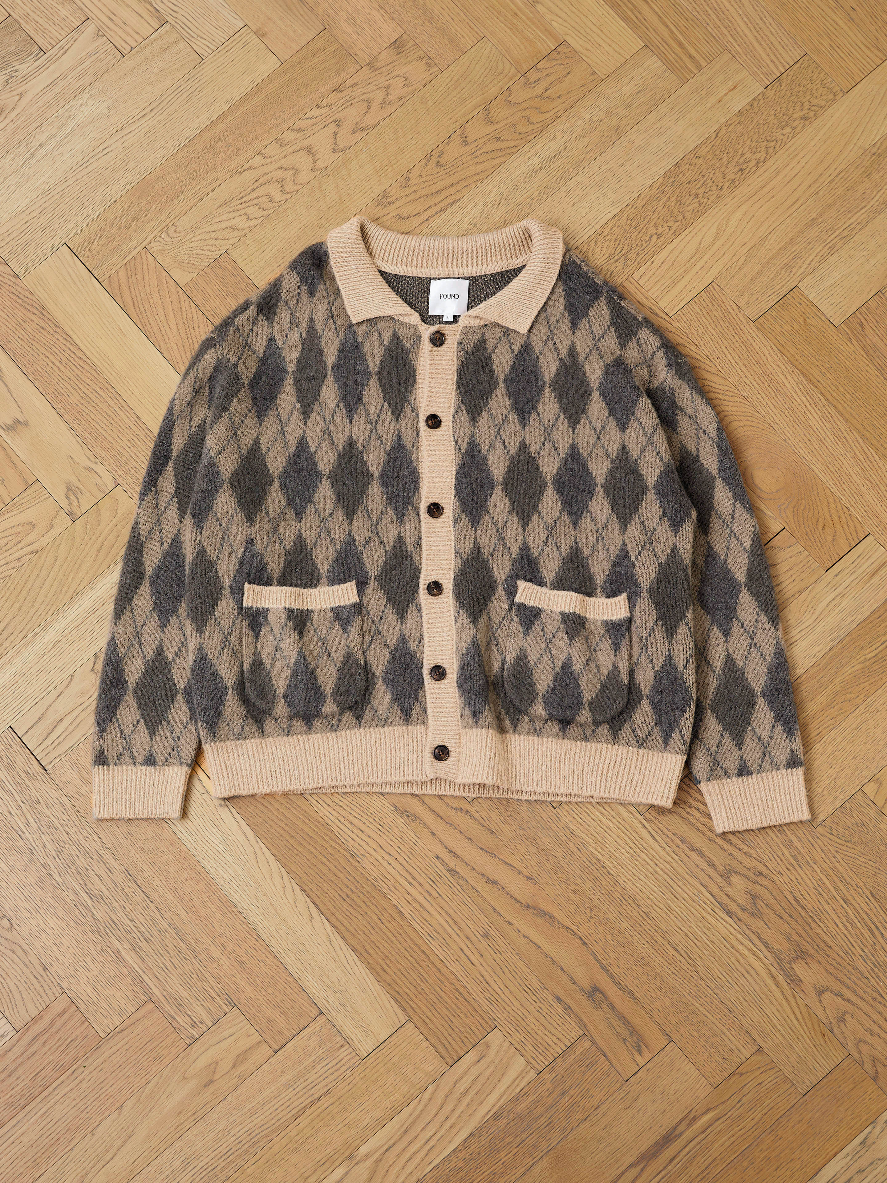 The FOUND Argyle Cardigan features a relaxed fit in brown and beige with black diamond patterns. Made from a cozy wool blend, it has a button-down front, two pockets, and ribbed cuffs and hem. Shown laid flat on a wooden herringbone floor.