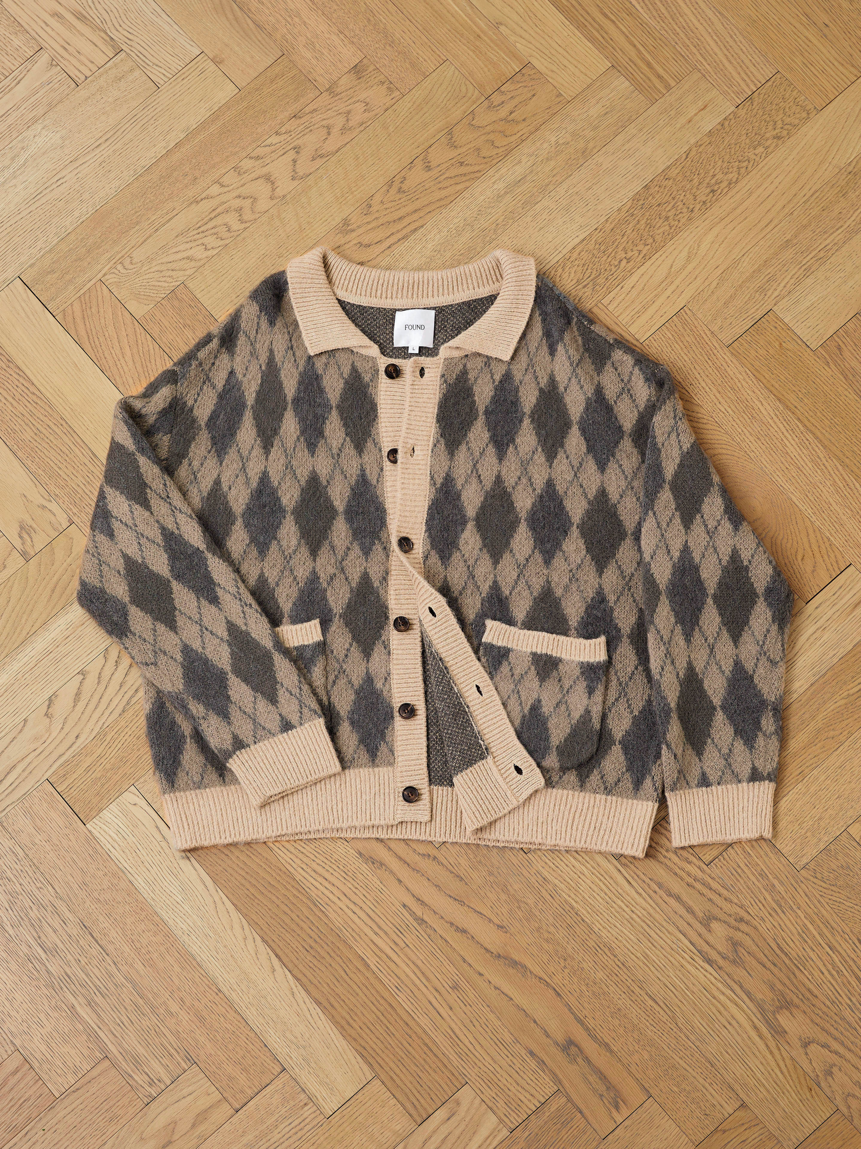 The FOUND Argyle Cardigan, crafted in a beige and gray wool blend with black buttons and two front pockets, is displayed flat on a wooden herringbone floor.