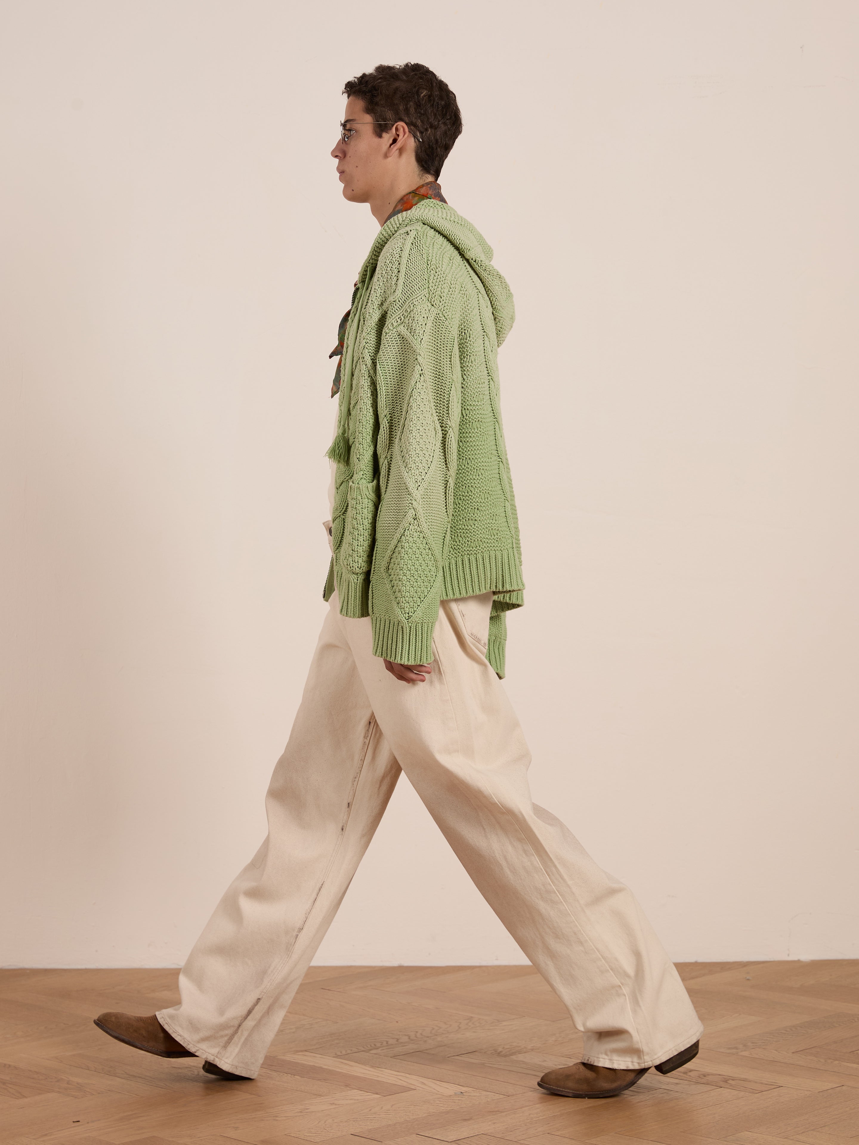 A person in a Zip Up Cable Knit Hoodie by FOUND, paired with beige wide-leg pants and brown shoes, walks on a wooden floor against a plain background.