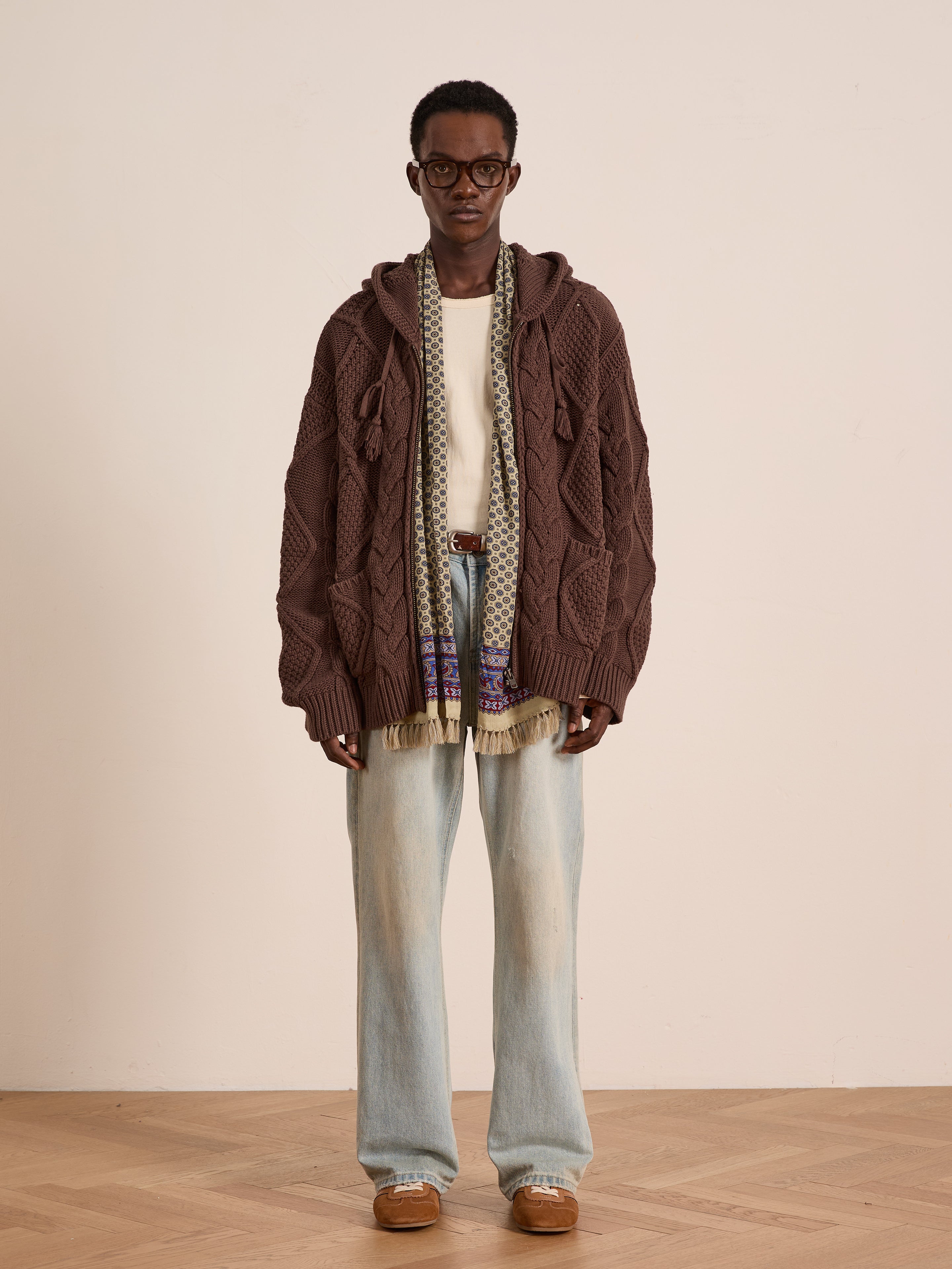 A person wearing glasses, the FOUND Zip Up Cable Knit Hoodie, a cream shirt, patterned scarf, light blue wide-leg jeans, and brown shoes stands against a plain light-colored wall.